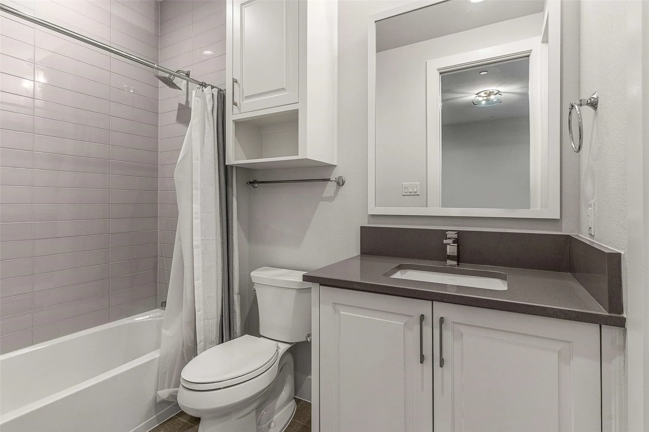 Contemporary bathroom featuring a white bathtub with light gray subway tile surround, a white vanity with shaker-style cabinetry, a dark countertop with an integrated sink, a framed mirror, and a built-in wall cabinet with open shelving