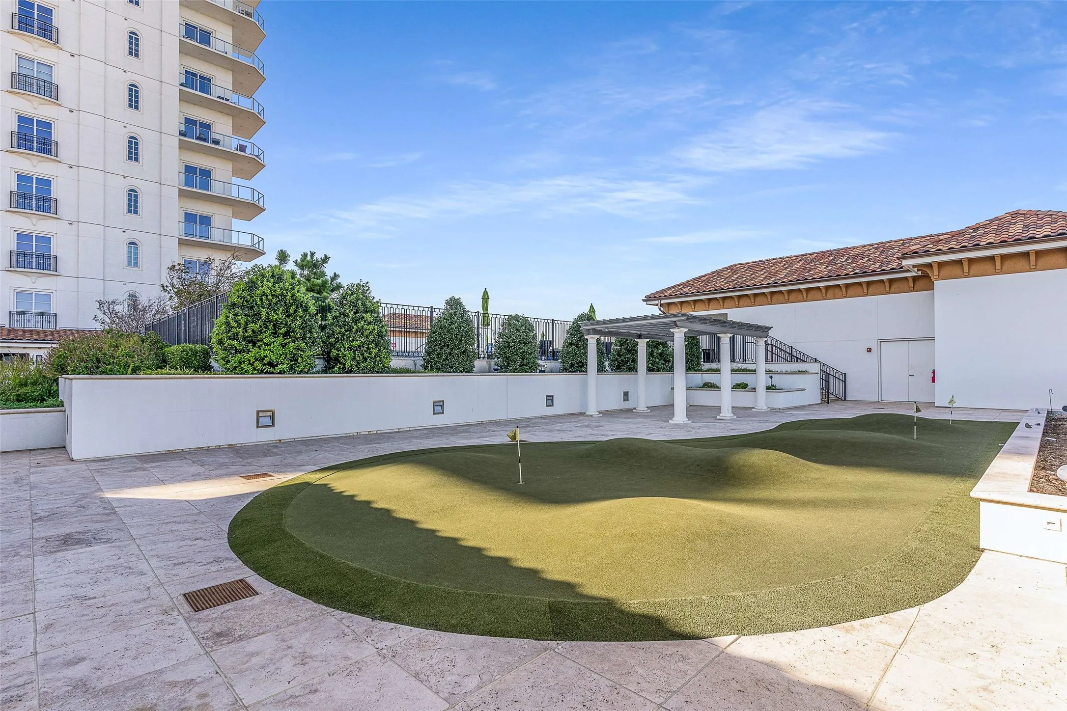 Outdoor putting green with undulating terrain, surrounded by a light-colored tiled patio, a white stucco building with a red tile roof, and a pergola with white columns