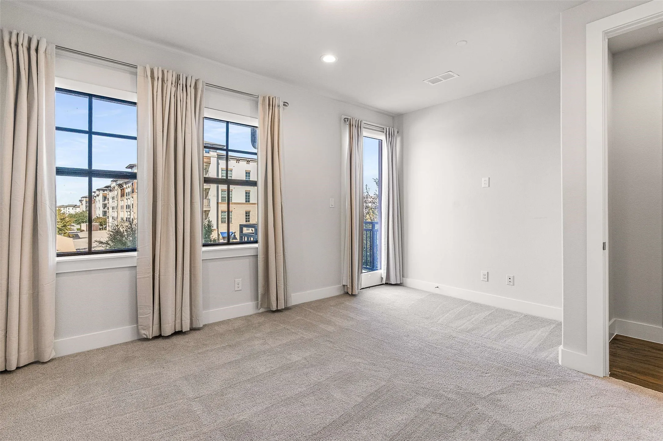 Carpeted room featuring two large windows with black framing, a glass-paneled door leading to a balcony with black railing, and white baseboards
