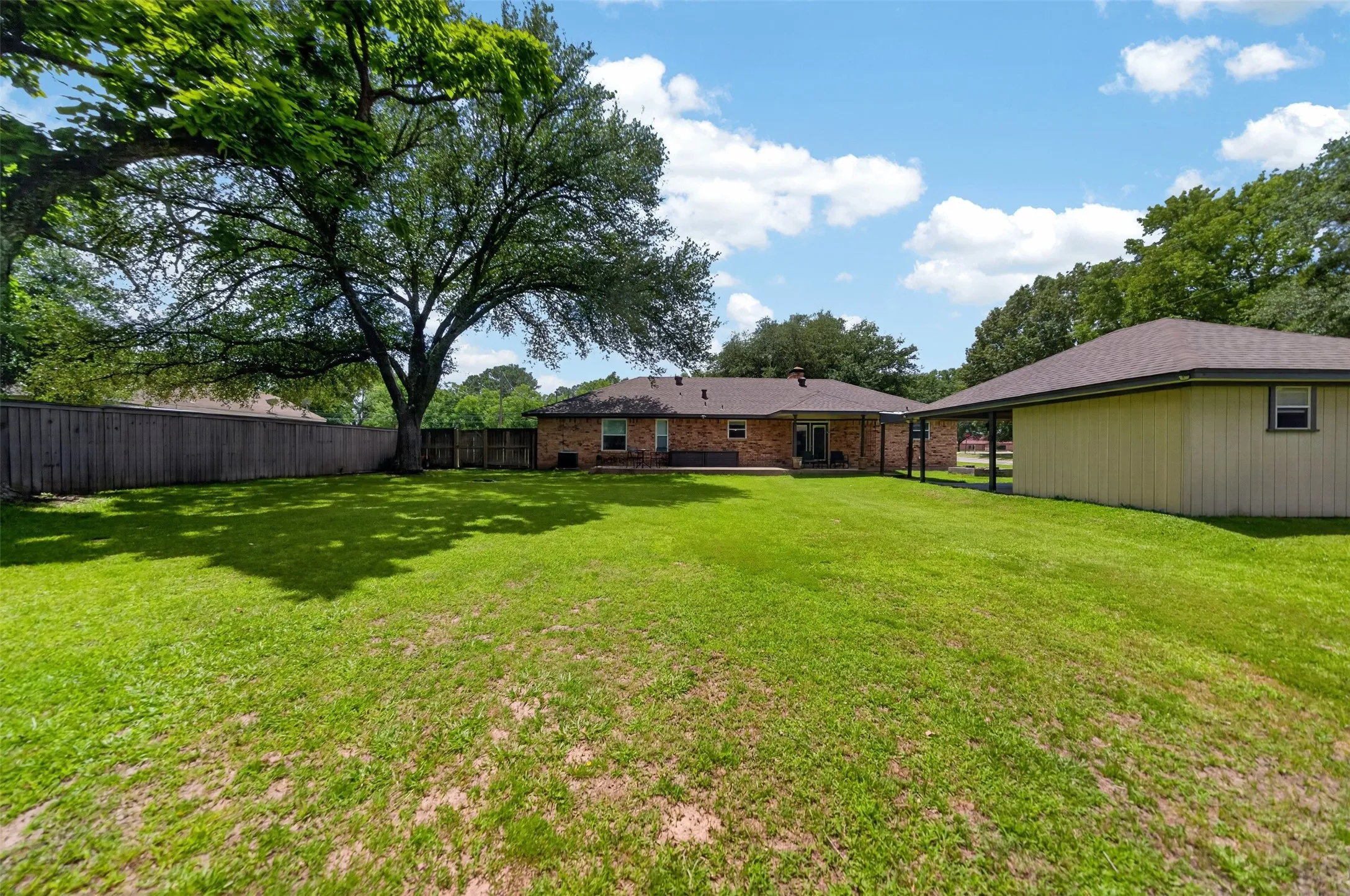 Expansive backyard featuring a mature shade tree, brick primary residence with a covered patio, separate auxiliary building with a covered walkway, and wood fencing enclosing the perimeter