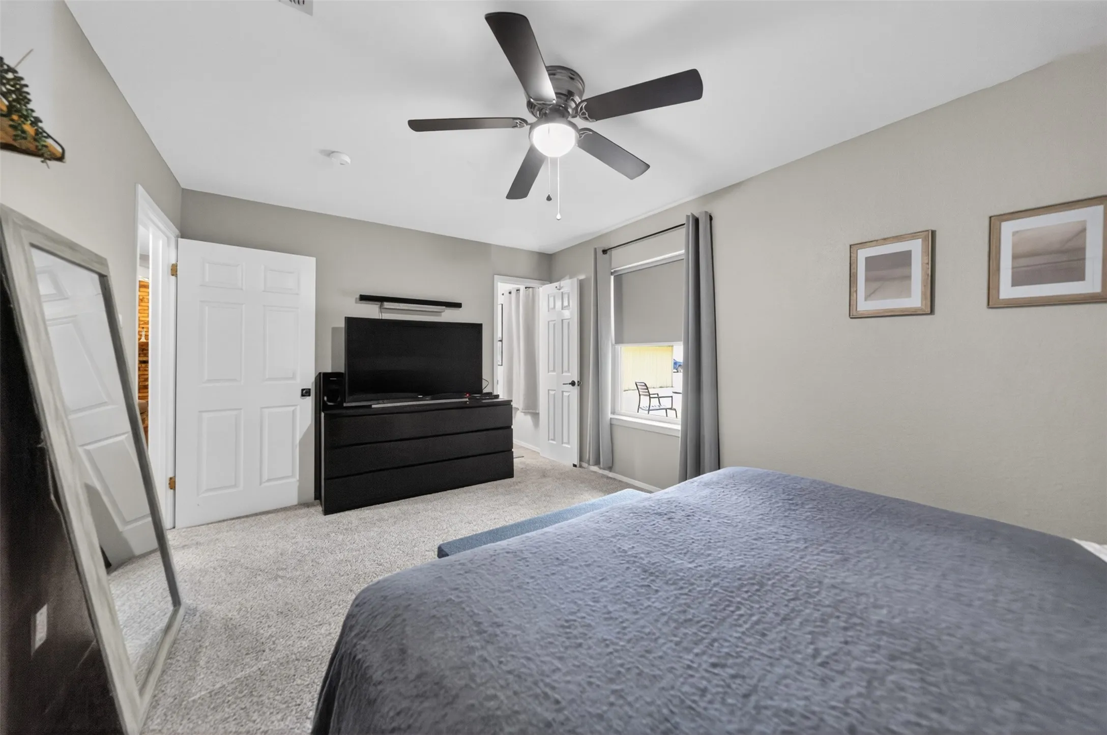Spacious interior room featuring light gray walls, light gray carpet flooring, a window with gray roller shade and curtains, and a ceiling fan with light fixture