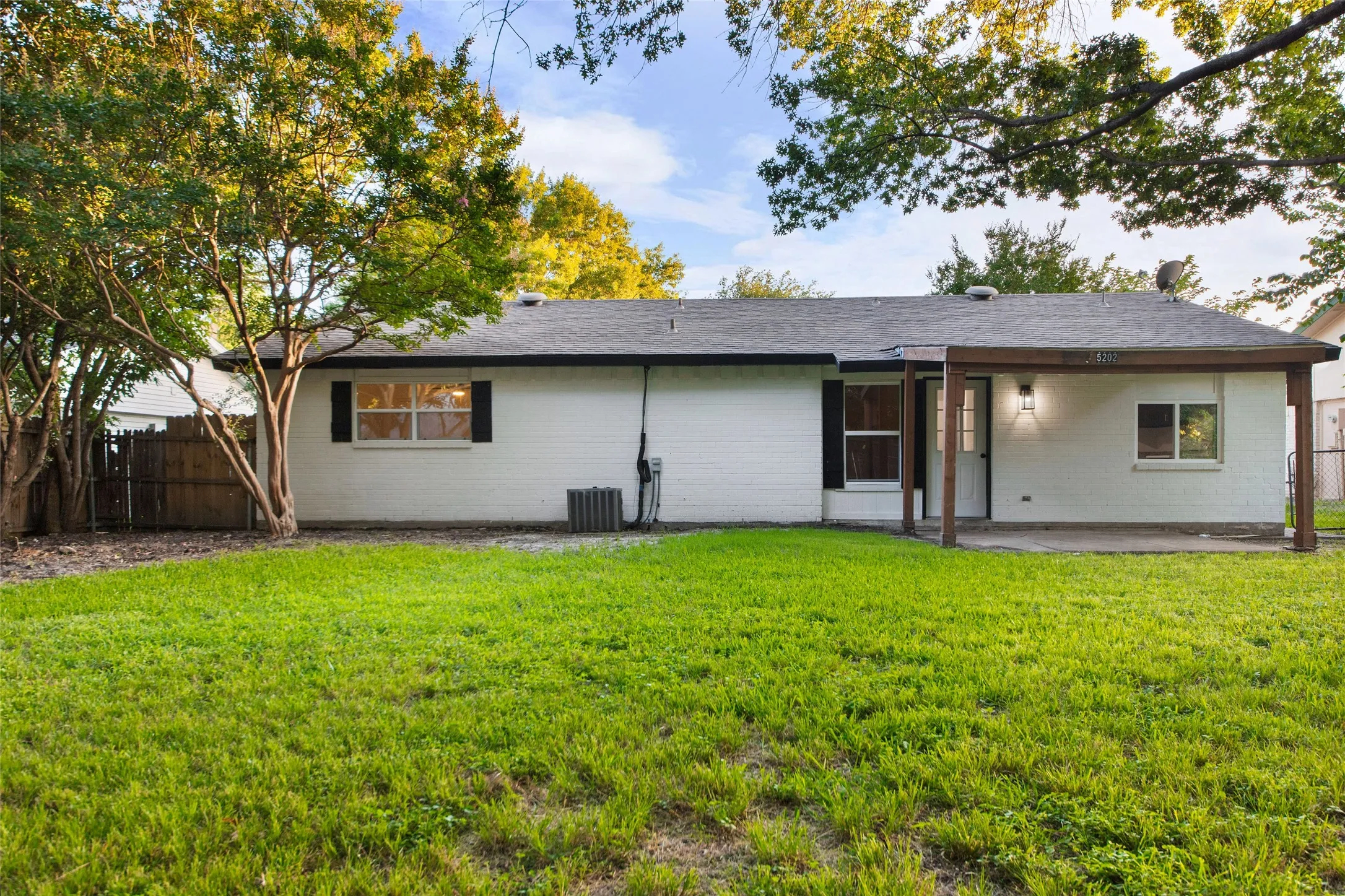 Expansive backyard featuring a covered patio, manicured lawn, mature trees, white exterior siding, and a dark roof
