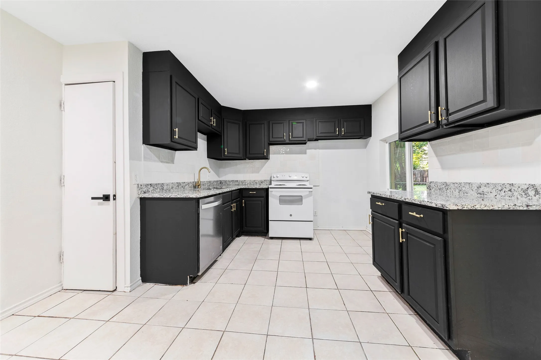 Kitchen featuring dark cabinetry with gold-tone hardware, a stainless steel dishwasher, a white range, light-toned tiled flooring, and a window with exterior views