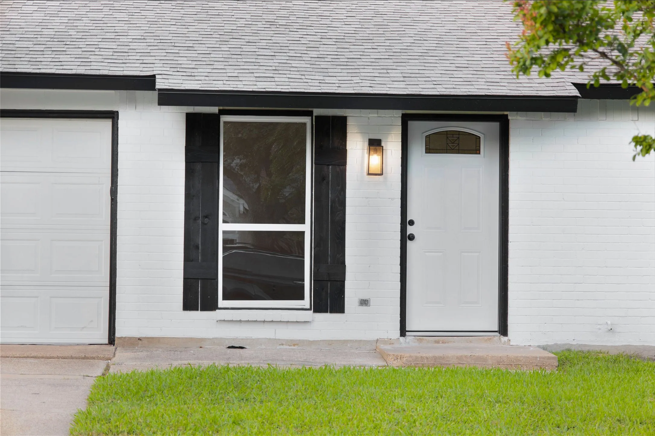 White painted brick exterior featuring a multi-paned front door with an arched glass insert, an outdoor sconce, a window with dark shutters, and a garage door