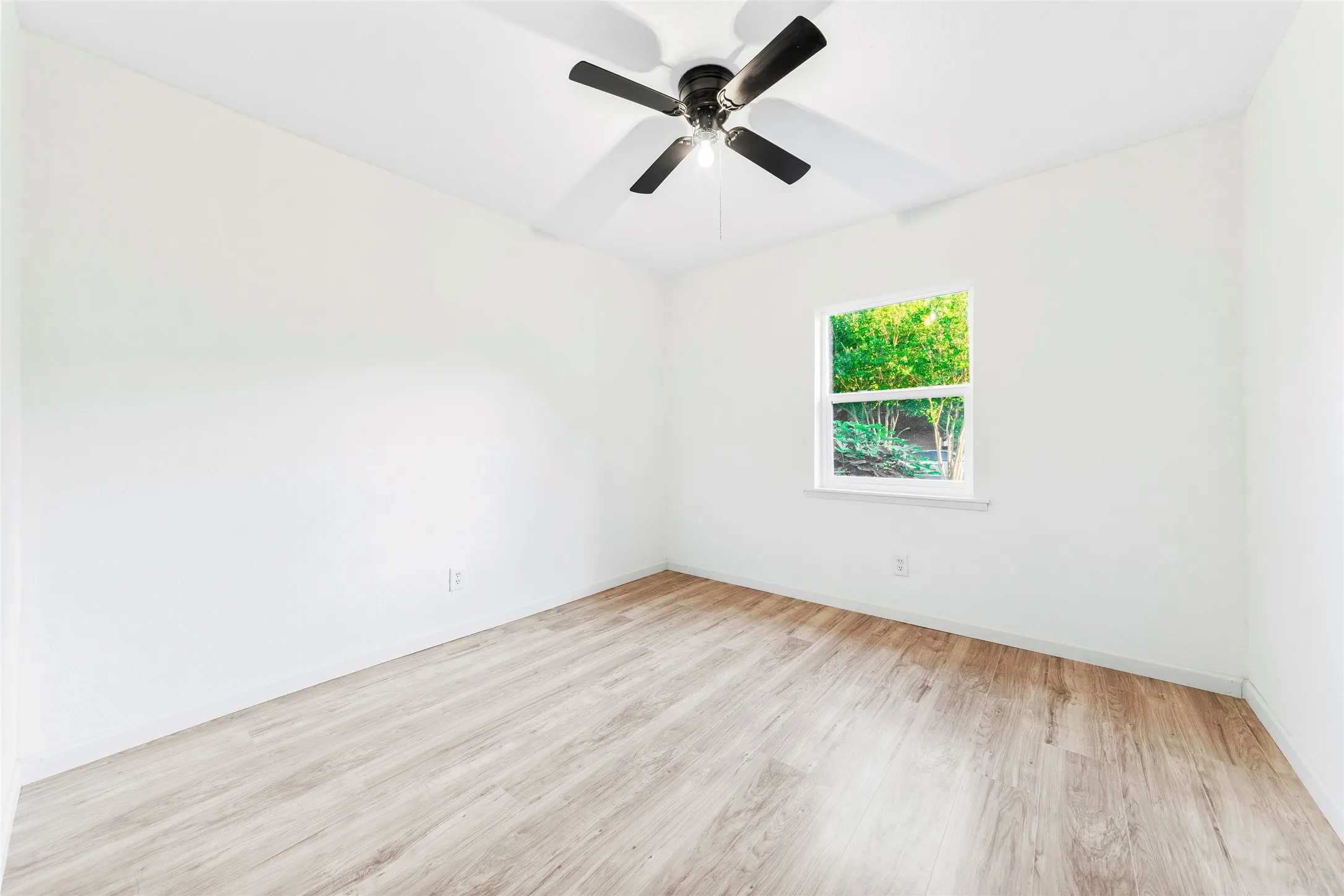 Room featuring wood-finish flooring, a contemporary ceiling fan with integrated lighting, and a single window offering exterior greenery