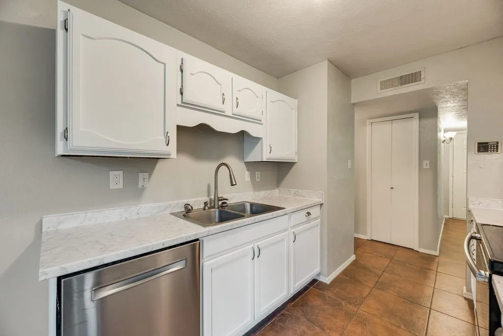 Kitchen featuring white cabinetry, light-toned countertops, a stainless steel dishwasher, a double basin sink with a gooseneck faucet, and brown tile flooring