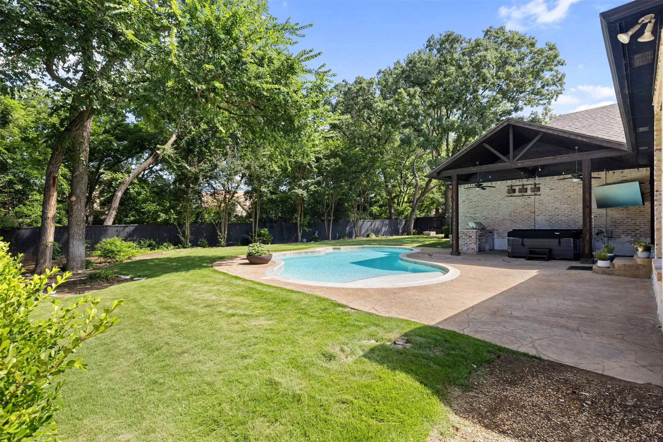 Backyard Oasis with pool, outdoor living center with built in grill, grass, trees, fence.