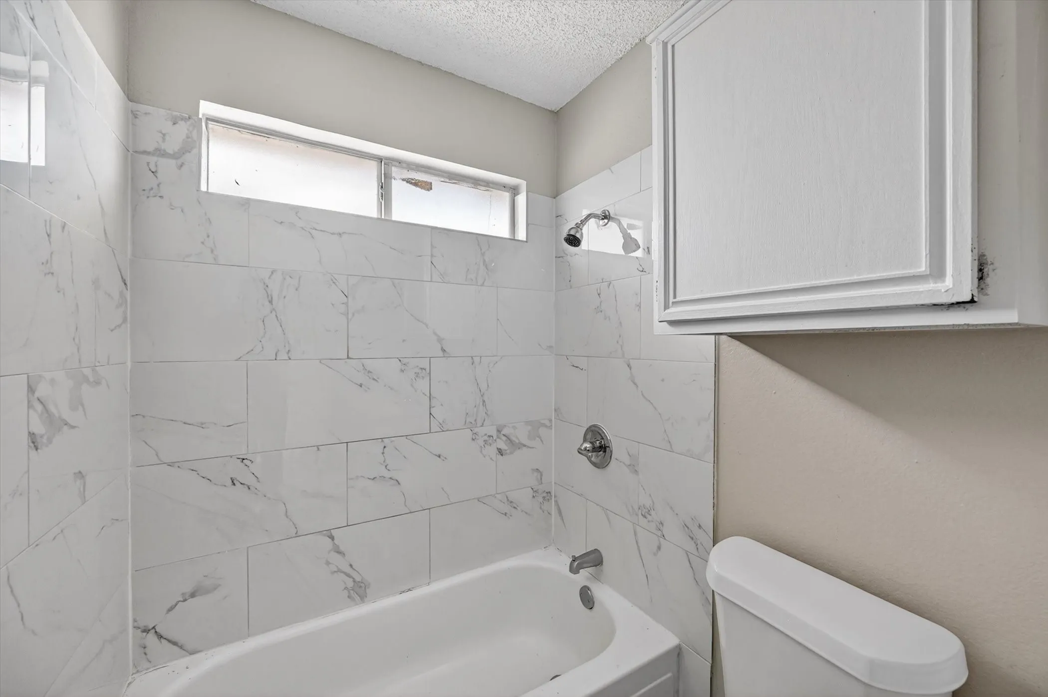 Bathtub with polished chrome fixtures, white tile surround with subtle gray veining, and an overhead transom window