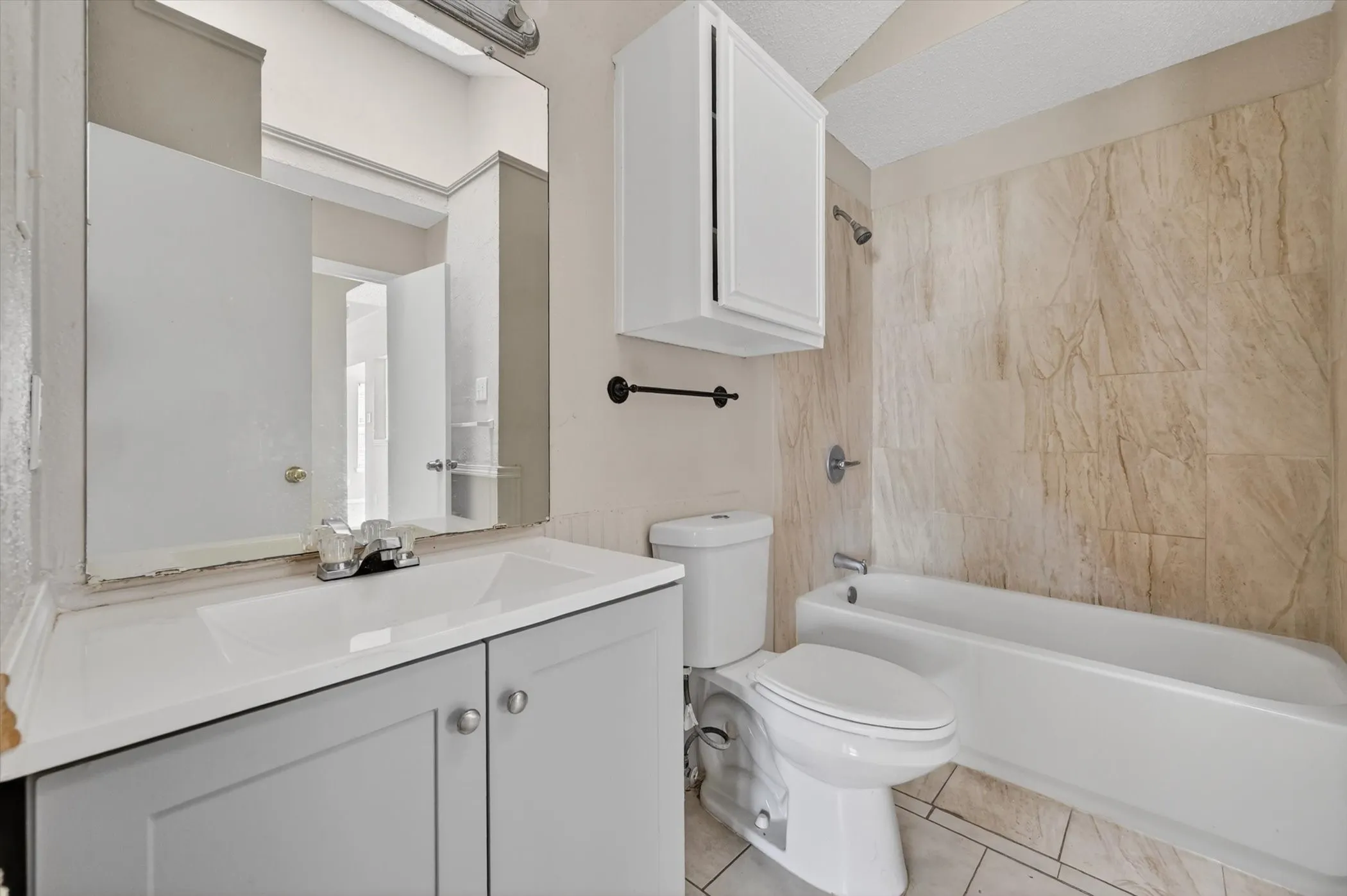 Bathroom featuring a single vanity with a white countertop and gray cabinetry, a wall-mounted mirror, a toilet, a bathtub, and natural-toned wall tiling
