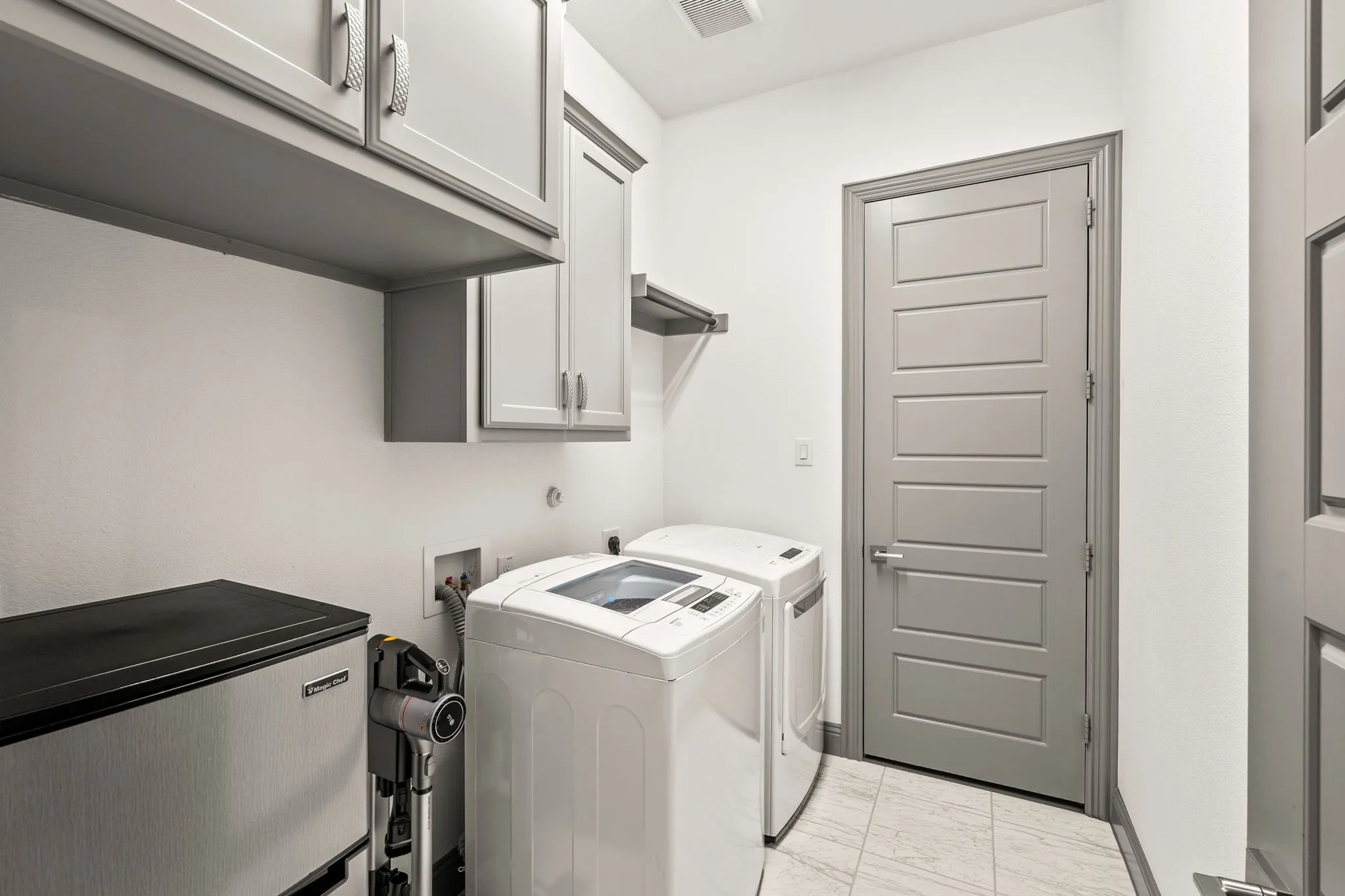 Laundry room featuring gray shaker-style cabinetry with chrome hardware, white walls, and light-toned tile flooring