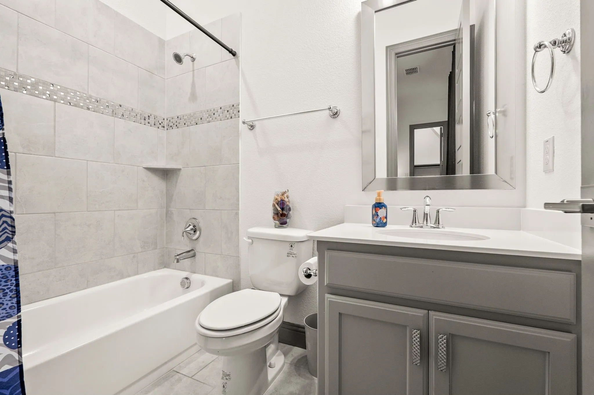 Bathroom featuring a tub/shower combination with tile surround and decorative accent band, a built-in shower niche, a single vanity with grey cabinetry, and a framed mirror
