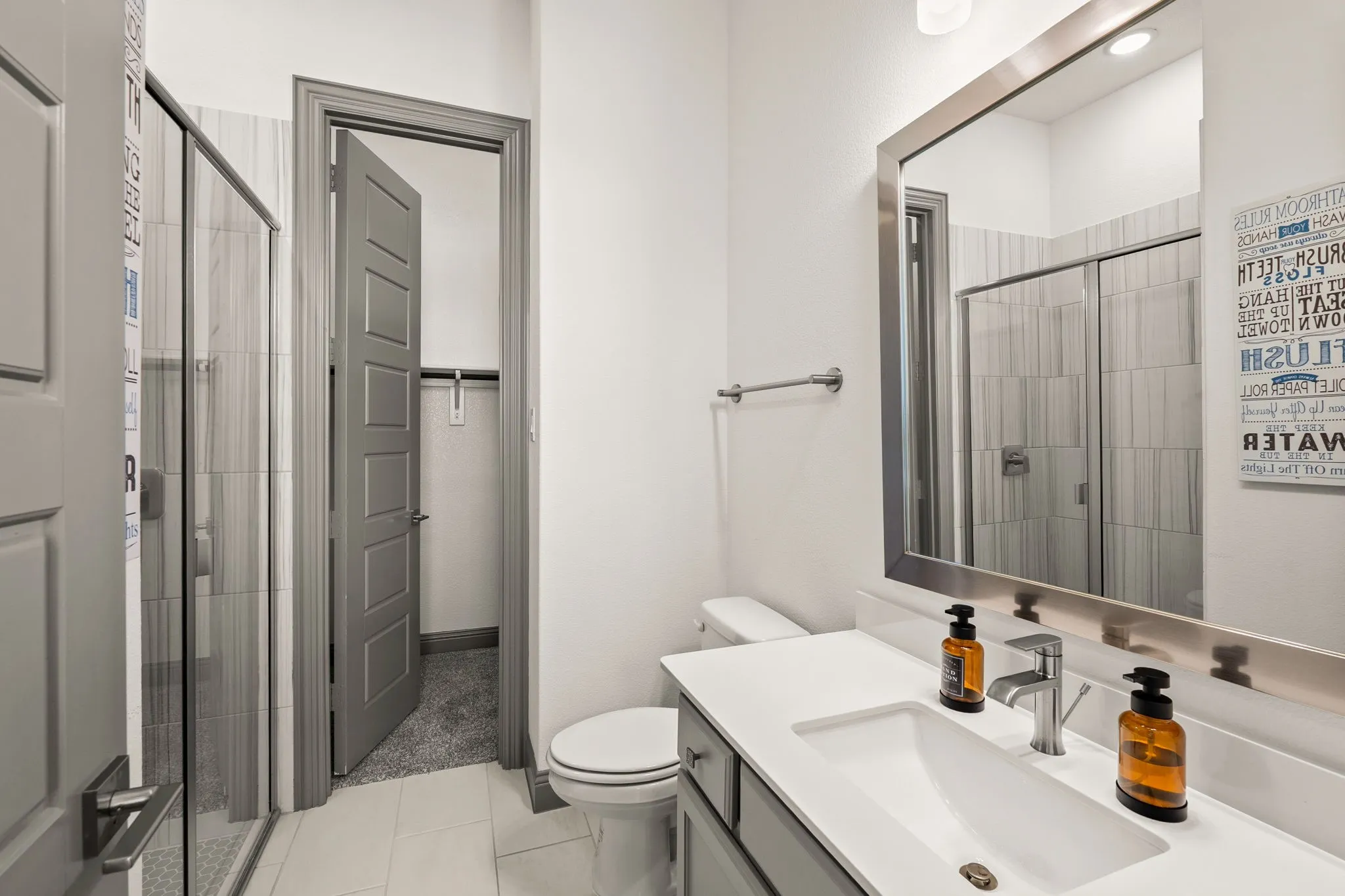 Bathroom featuring a shower with glass enclosure, white rectangular vanity sink, gray paneled vanity cabinetry, full-width mirror with metal frame, and light-colored floor tiles