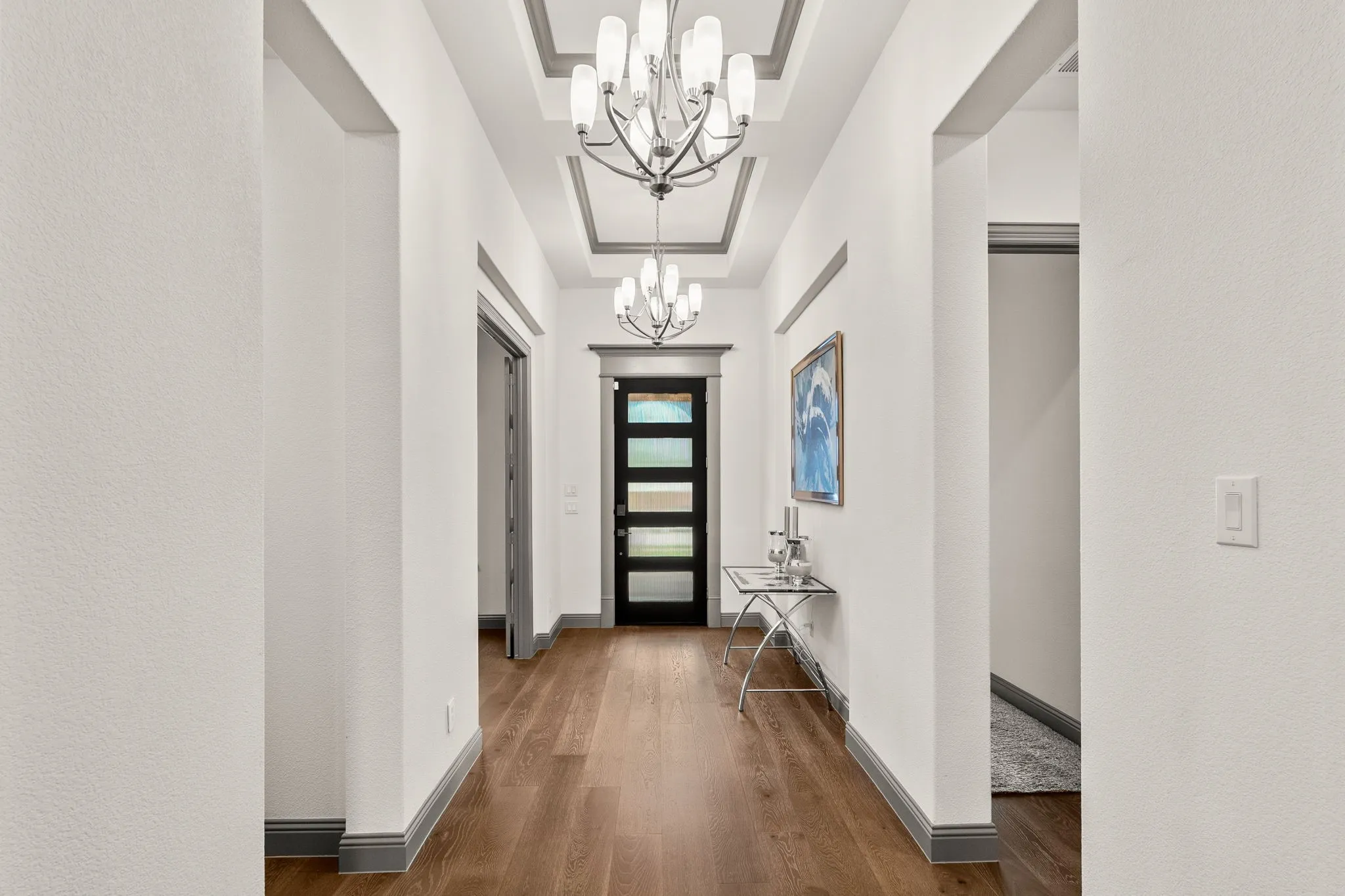Entryway featuring wood-finish flooring, a modern black door with frosted glass panels, and a coffered ceiling with two contemporary chandeliers