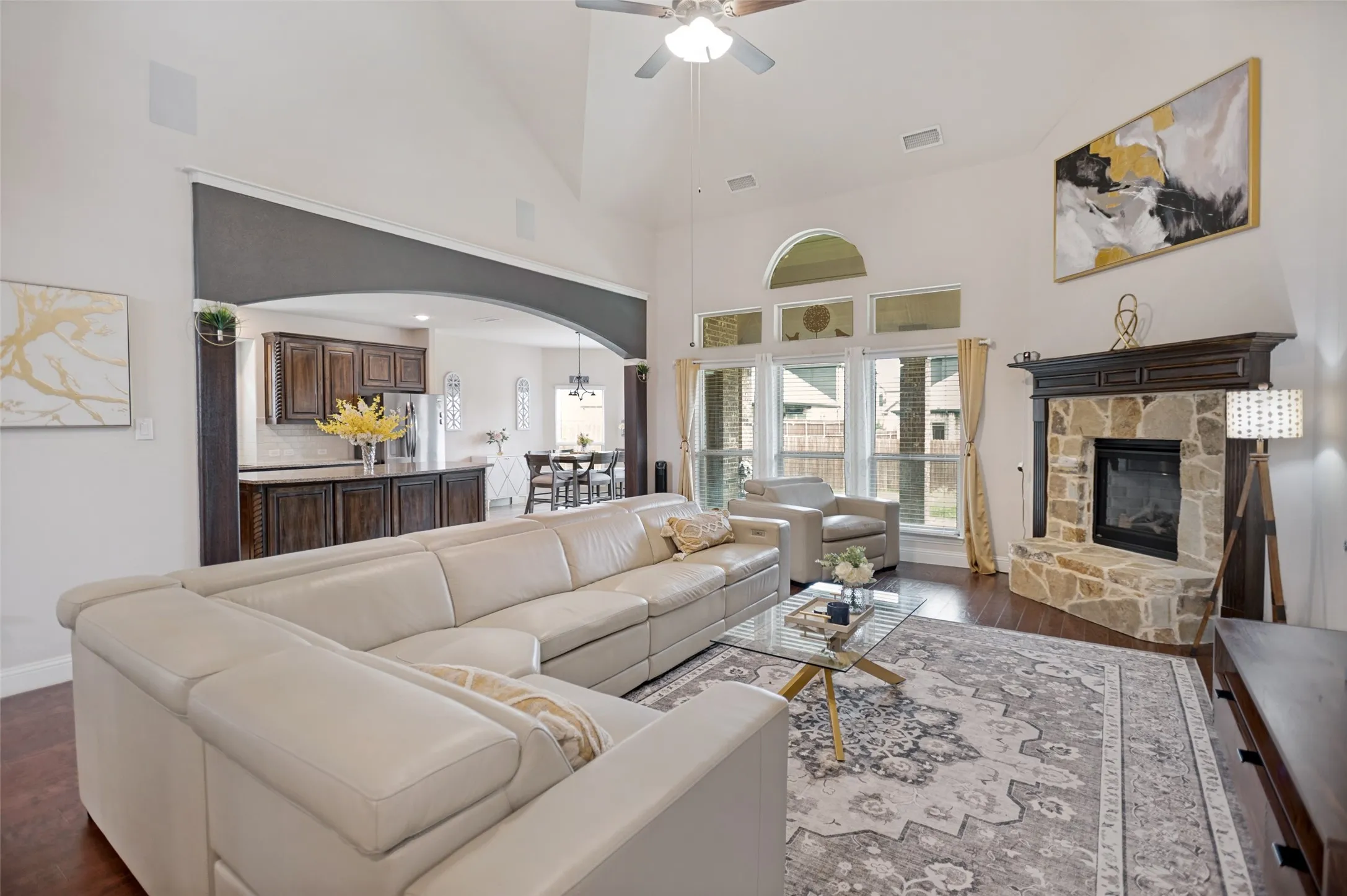 Living area featuring vaulted ceilings, an arched entryway to the kitchen, wood-finish flooring, a stone fireplace with a dark wood mantel, and large windows with an arched transom