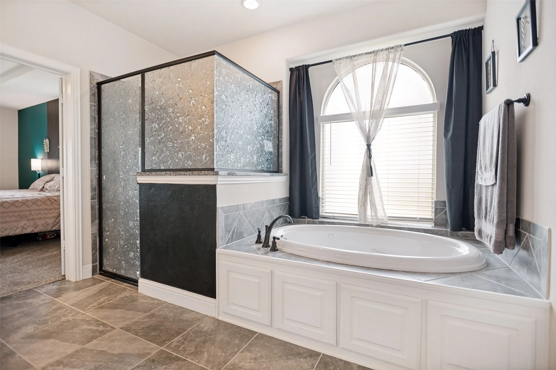 Primary bathroom with soaking tub set against an arched window, complemented by a textured glass shower enclosure with a dark frame, and finished with large format tile flooring and wainscoting