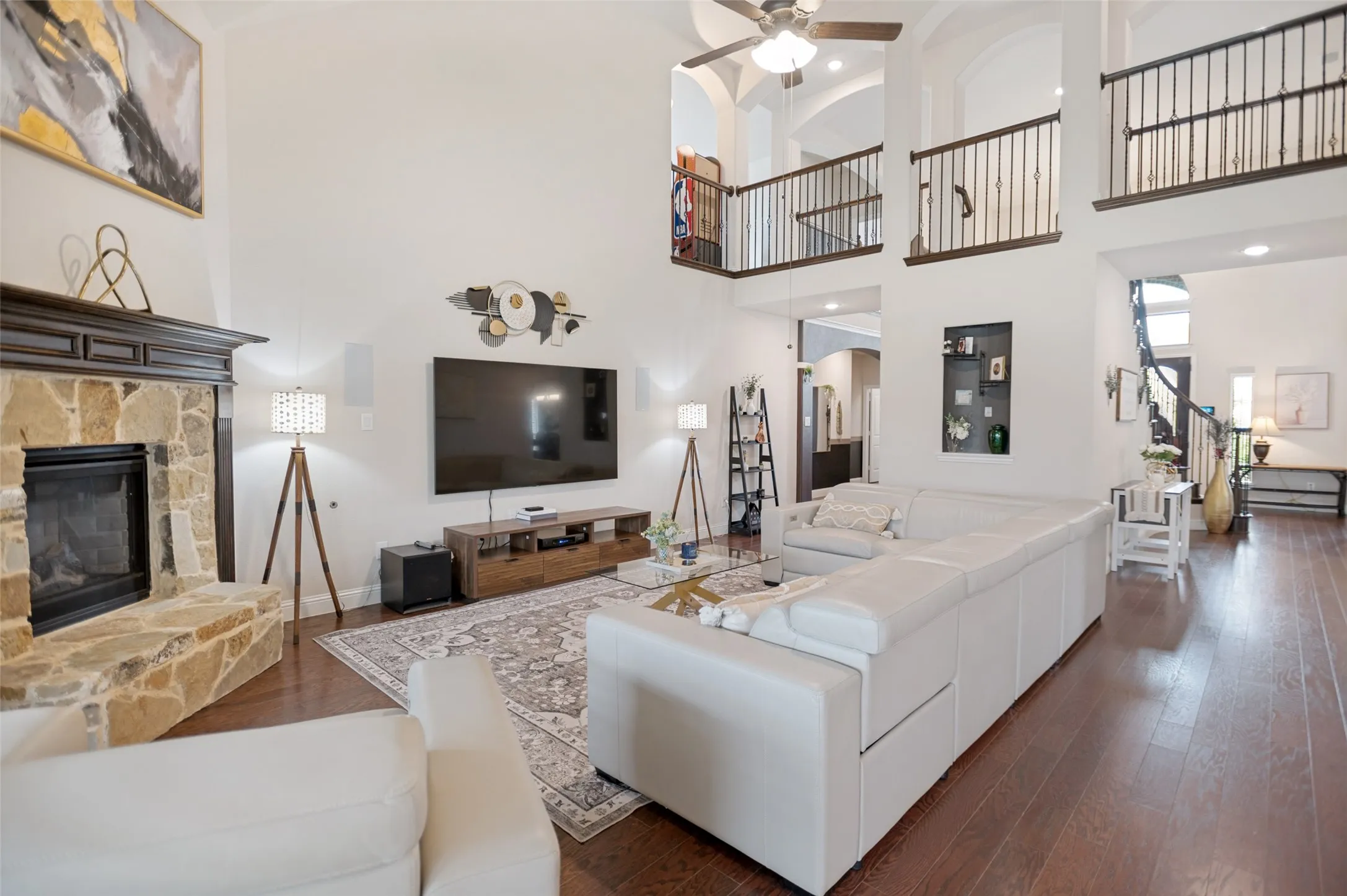 Two-story living area with high ceilings, featuring a stone-clad fireplace with a dark wood mantel, wood-finish flooring, and a second-story balcony with metal railings
