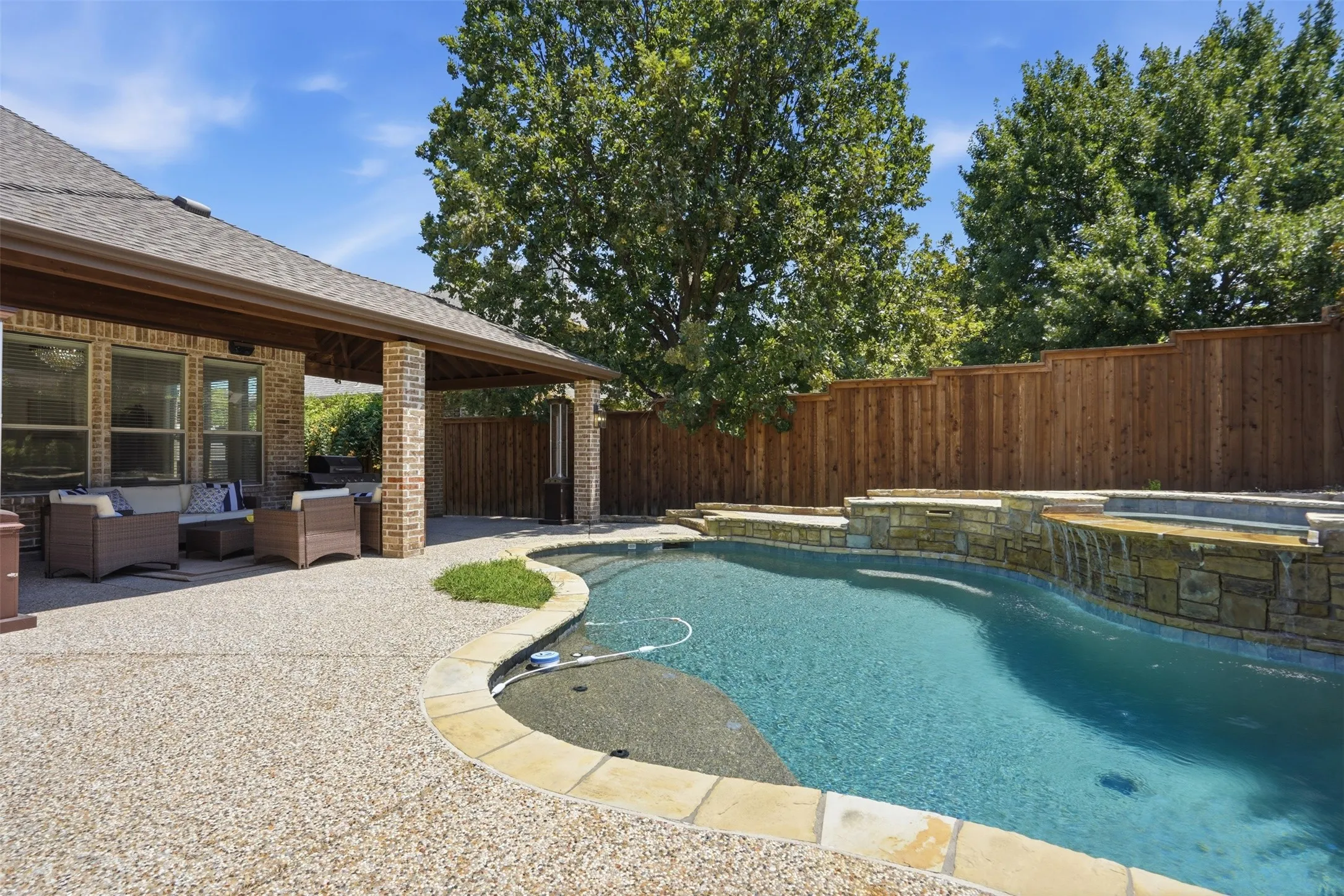Expansive outdoor living area featuring a covered patio, exposed aggregate concrete deck, and a curved swimming pool with a stone-clad water feature