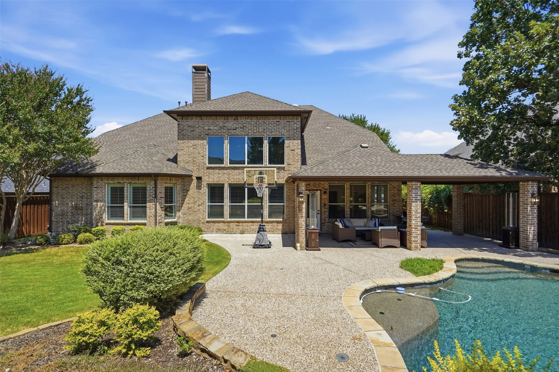 Brick exterior with multiple windows, covered patio, textured concrete patio, and a curved inground pool