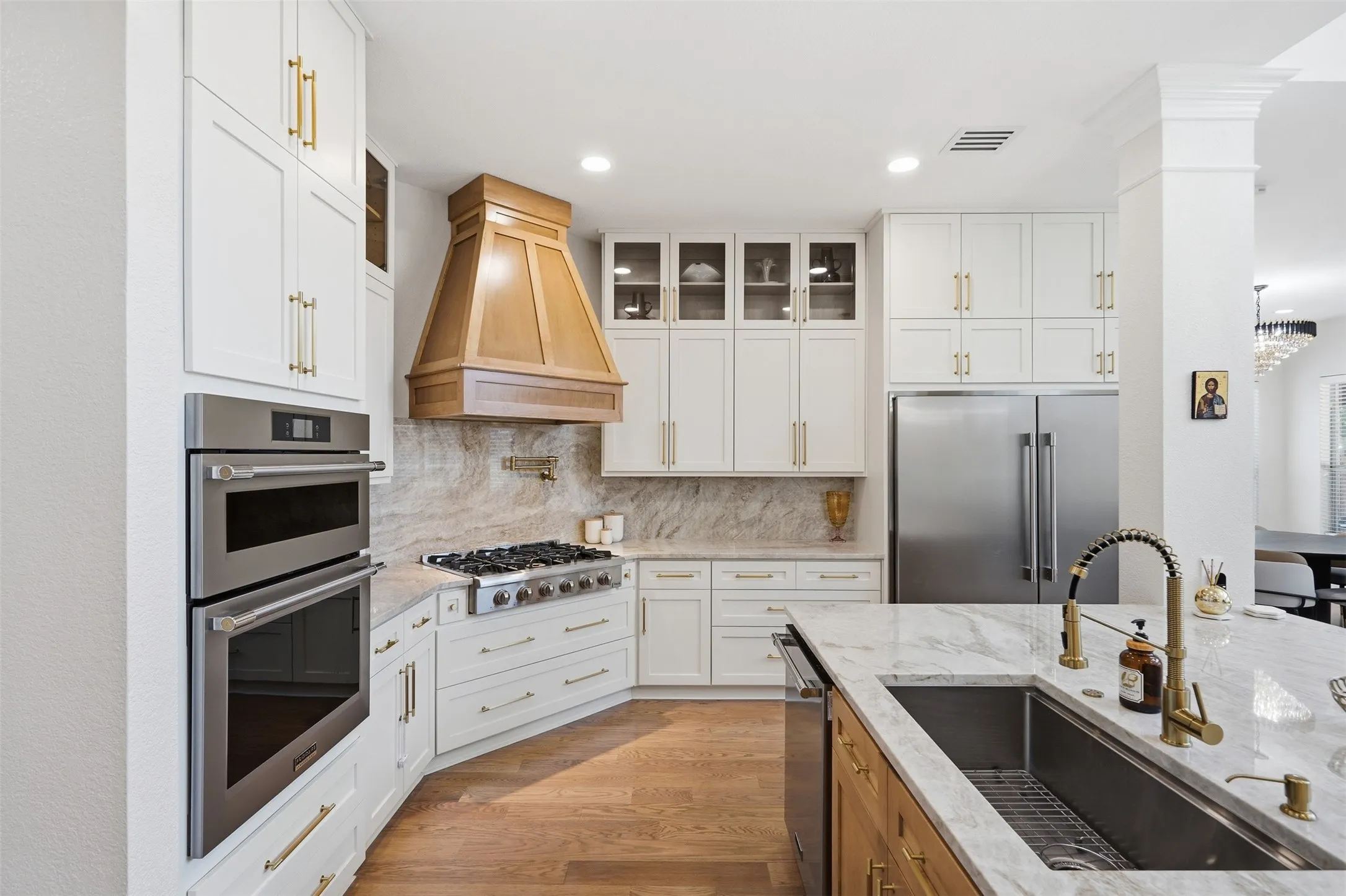 Chef's kitchen featuring white cabinetry with brass hardware, a copper range hood, dual wall ovens, stainless steel refrigerator, gas cooktop, and marble-finish countertops with waterfall island edge