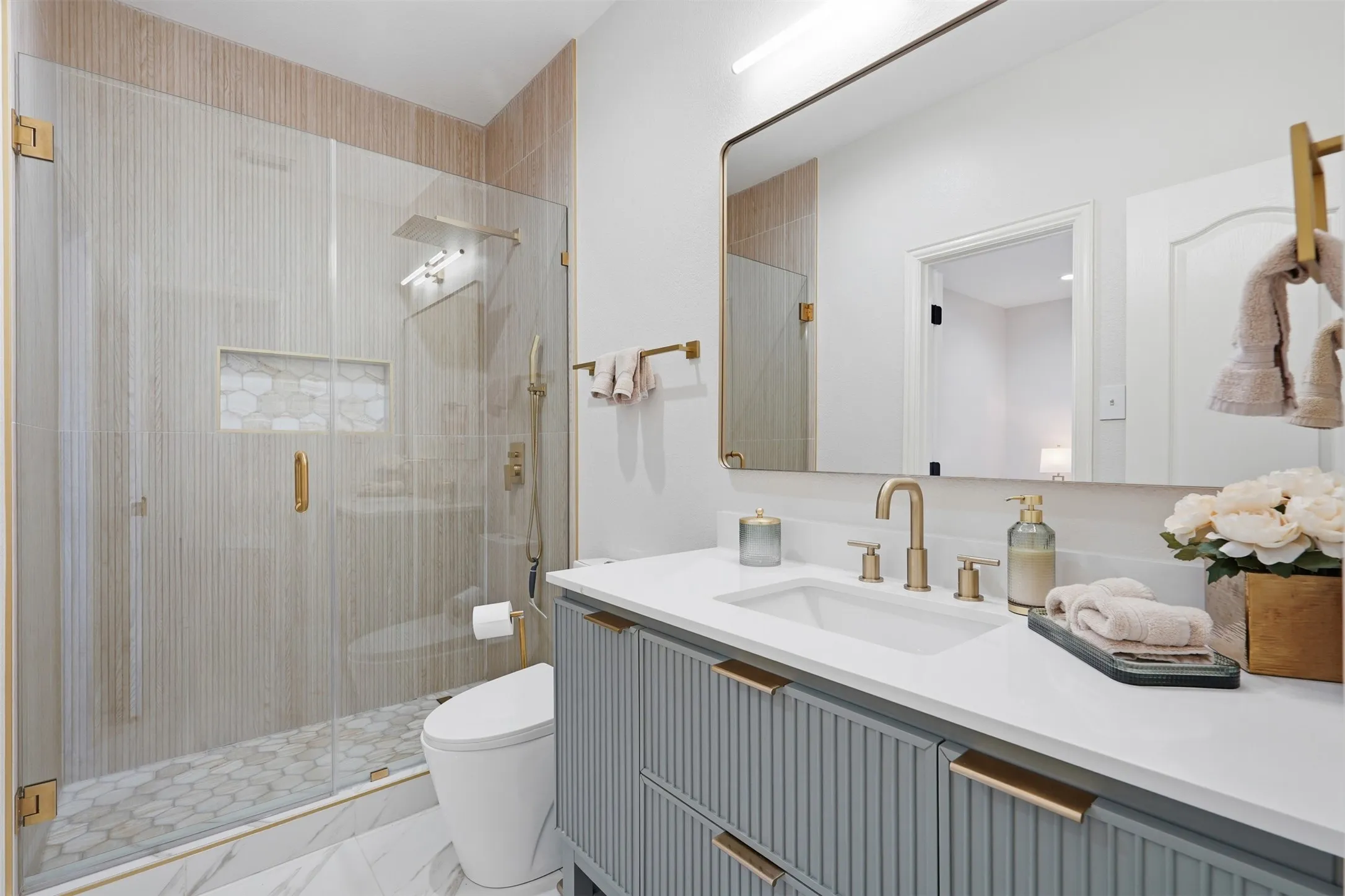 Glass-enclosed shower with a tiled niche and hexagonal floor, complemented by a single vanity with a white countertop and gold-toned hardware