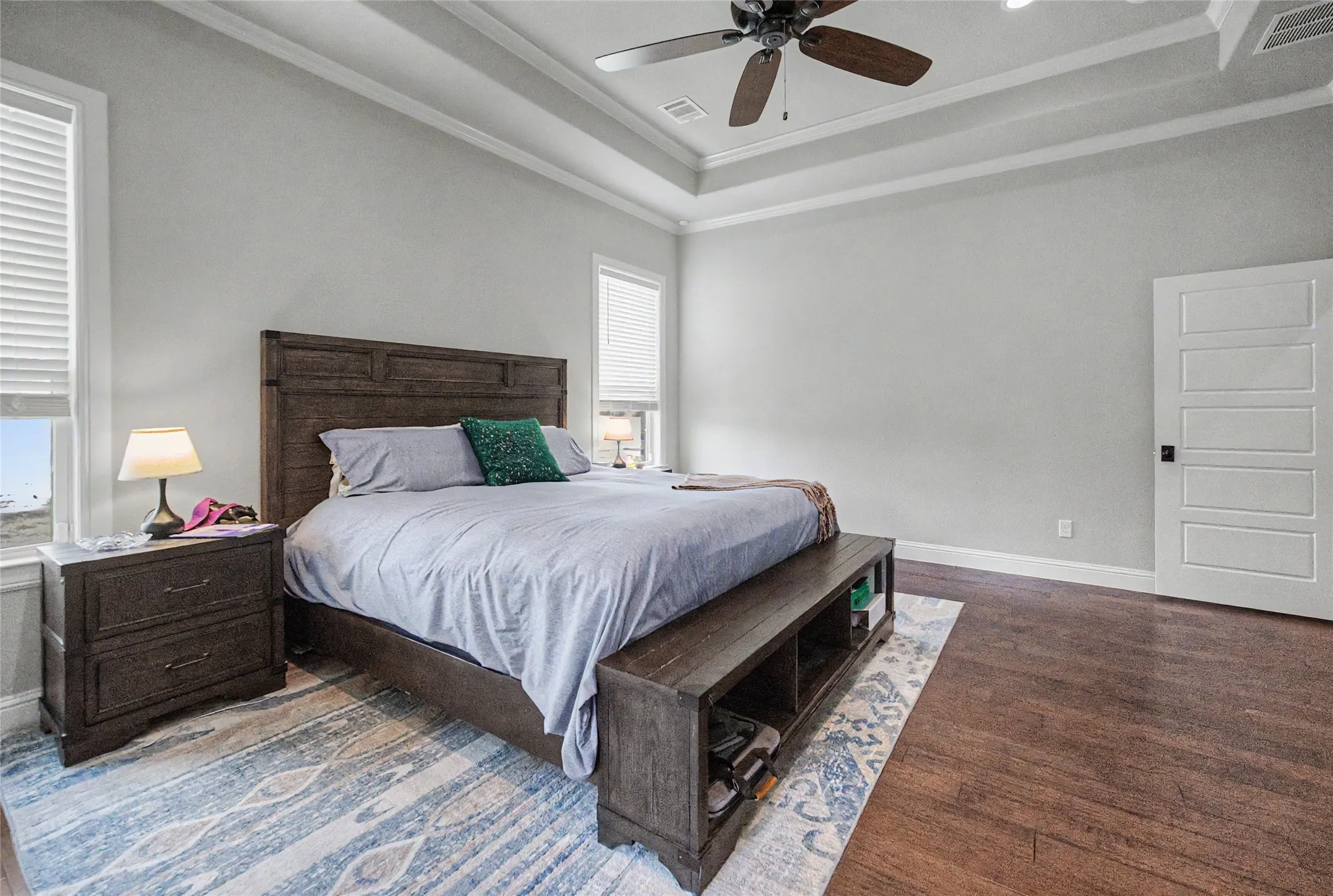 Recessed tray ceiling with crown molding, ceiling fan, wood-finish flooring, two windows with blinds, and a five-panel interior door