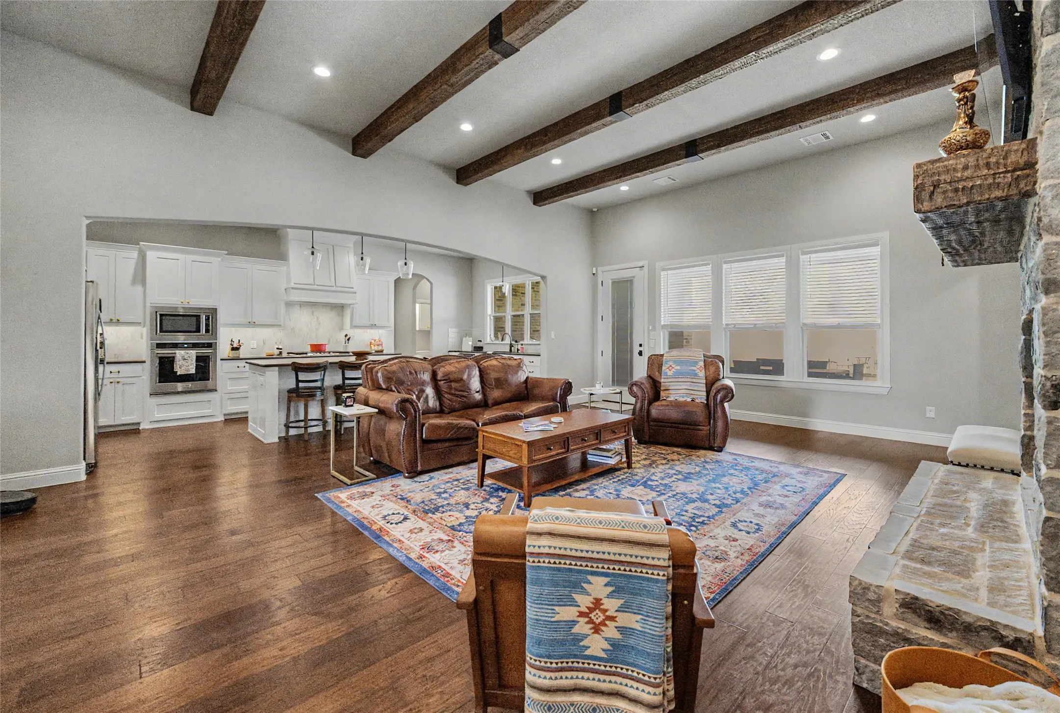 Open-concept living space featuring vaulted ceilings with exposed wood beams, wood-finish flooring, a stone fireplace, and multiple windows