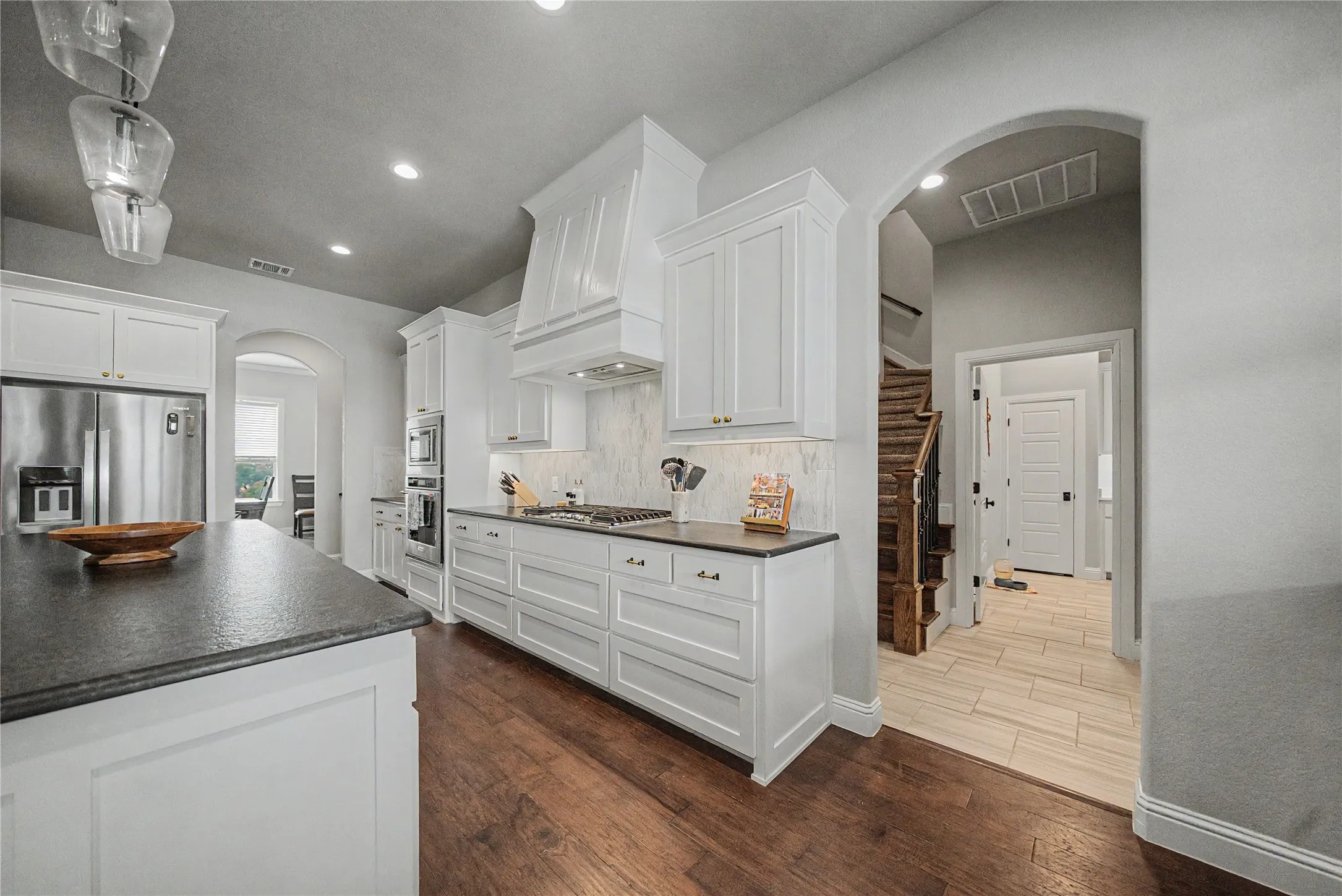 Kitchen featuring white cabinetry, dark stone countertops, stainless steel appliances, wood-finish flooring, and a decorative range hood