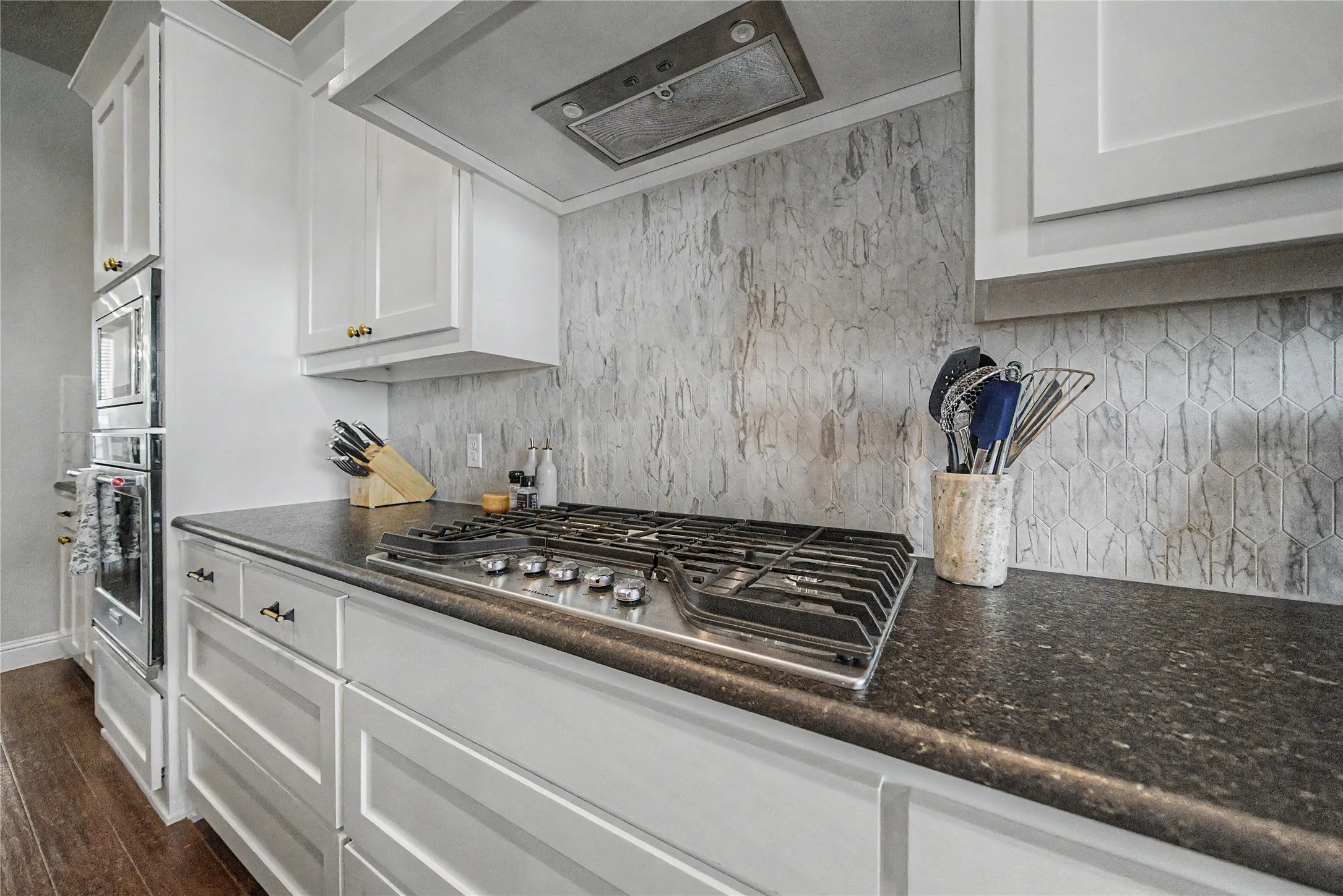 Kitchen featuring a gas cooktop, built-in double ovens, white cabinetry with gold-tone hardware, dark countertops, and a patterned backsplash