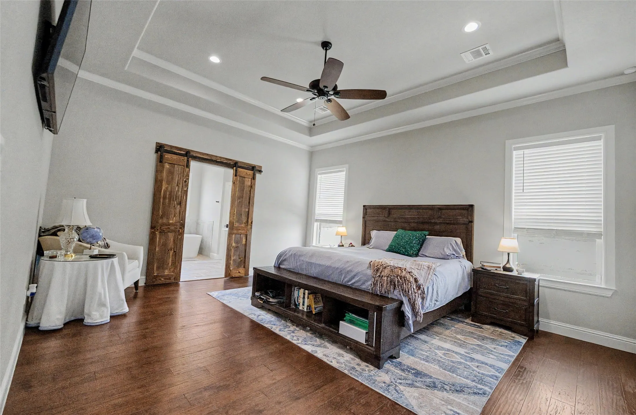 Spacious bedroom with rich wood-finish flooring, two large windows with blinds, and a tray ceiling featuring a ceiling fan and recessed lighting