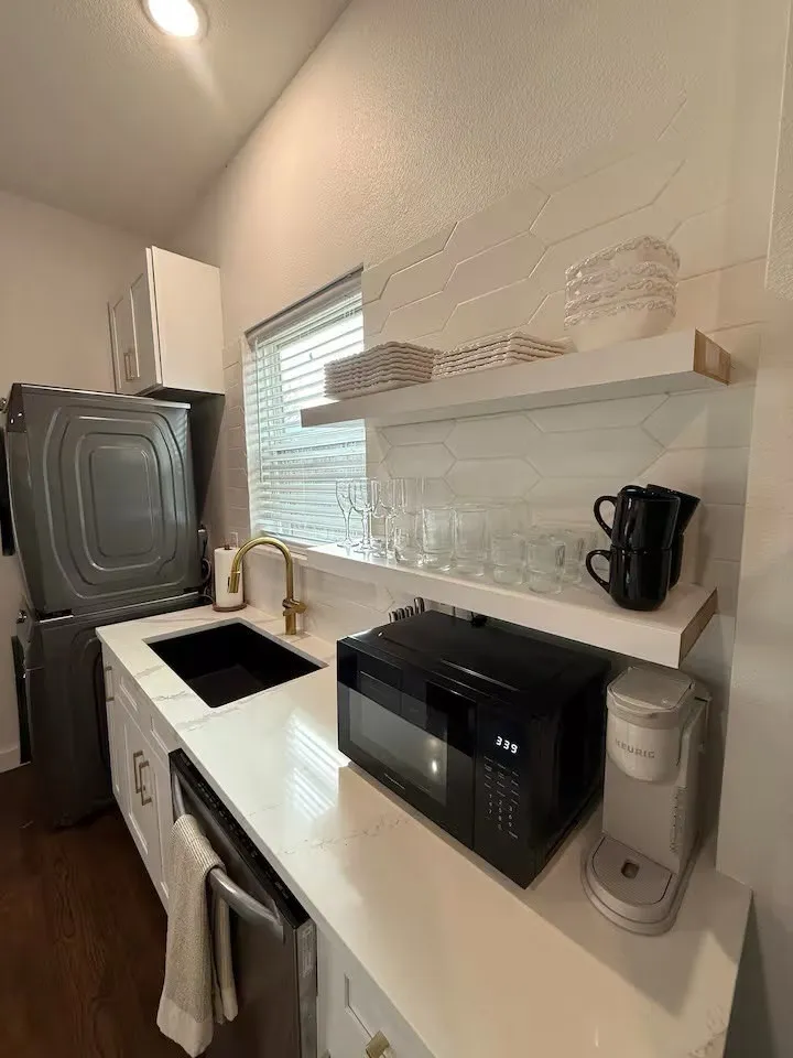 Compact kitchen area featuring a white hexagonal tile backsplash, white floating shelves, and a white countertop with an integrated black sink
