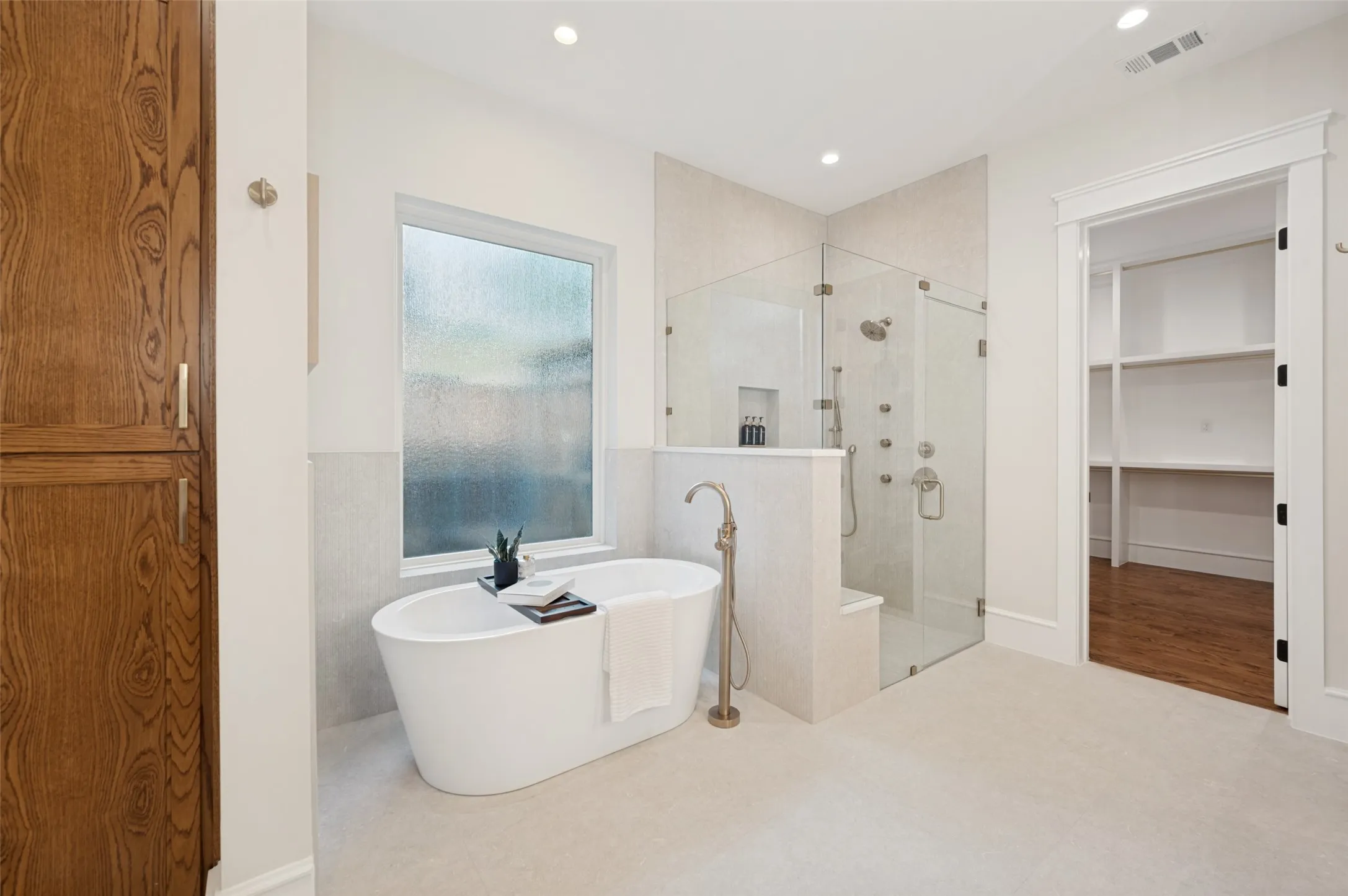 Freestanding soaking tub with floor-mounted faucet, oversized glass-enclosed shower with multiple sprayers, wood-paneled built-in storage cabinetry, and recessed lighting