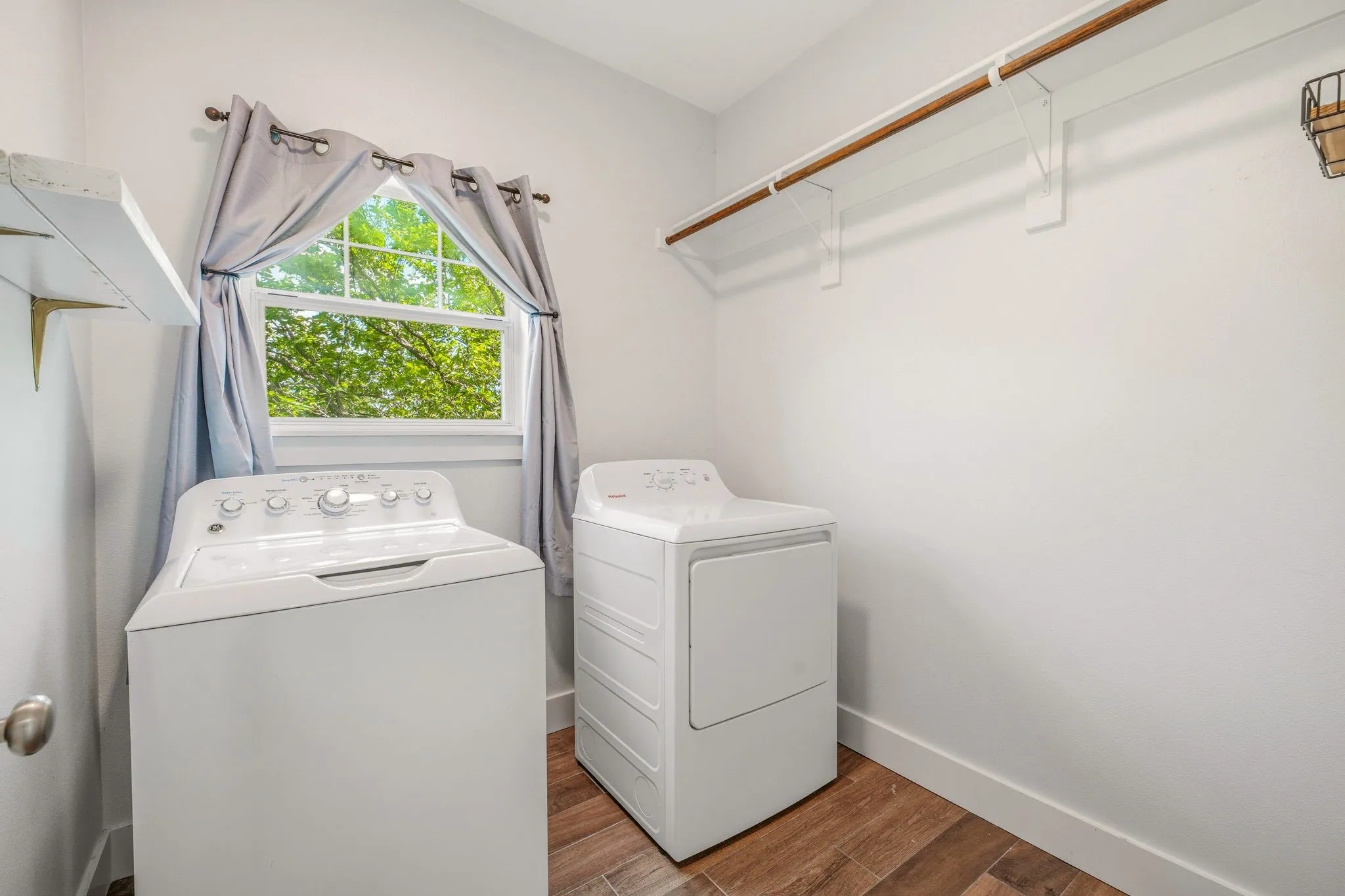 Dedicated laundry area featuring wood-finish flooring, a white window frame, and a wall-mounted wooden hanging rod with shelving