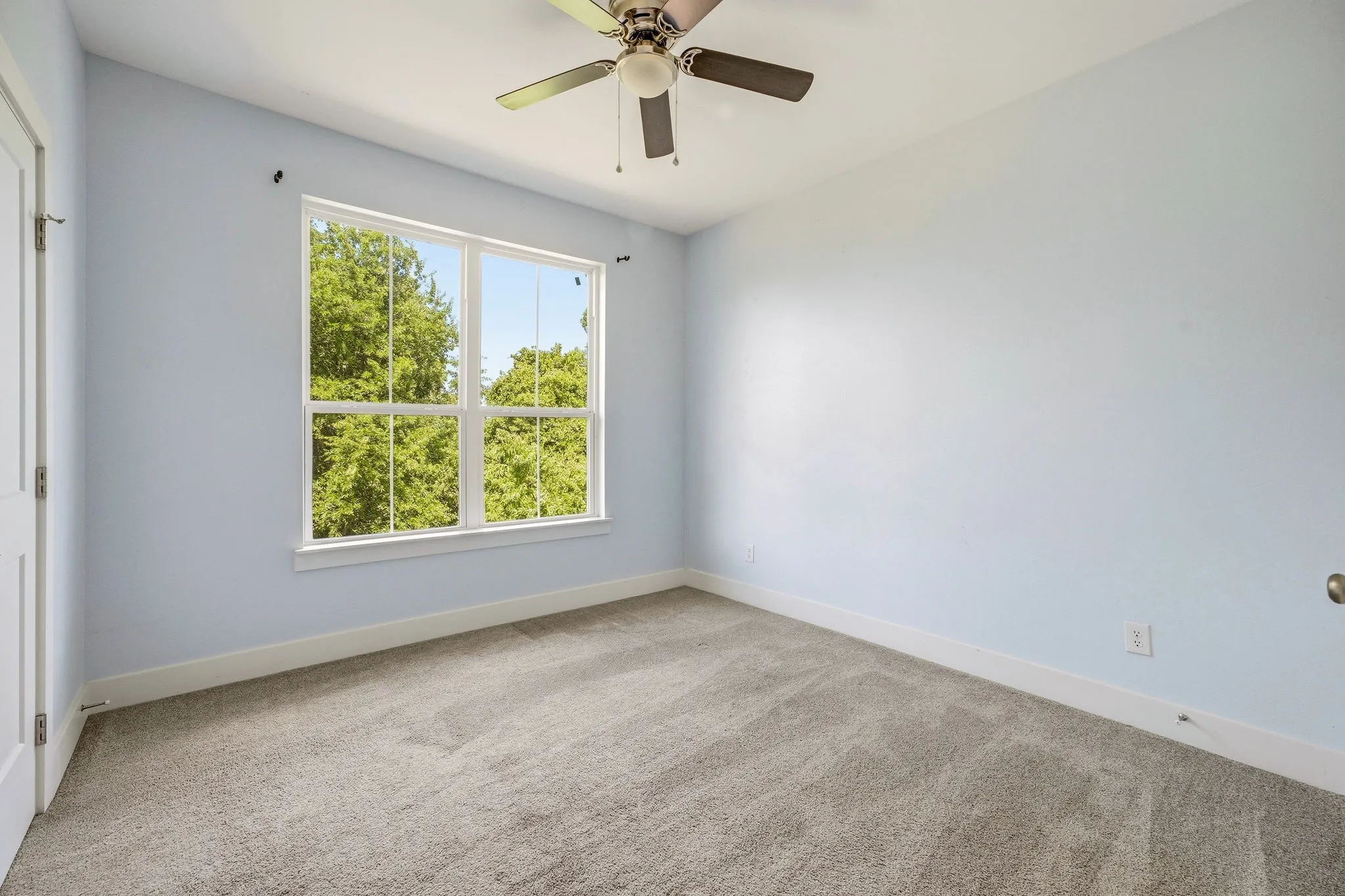 Spacious room featuring a large window with white trim, light blue wall color, gray carpet, white baseboards, and a ceiling fan