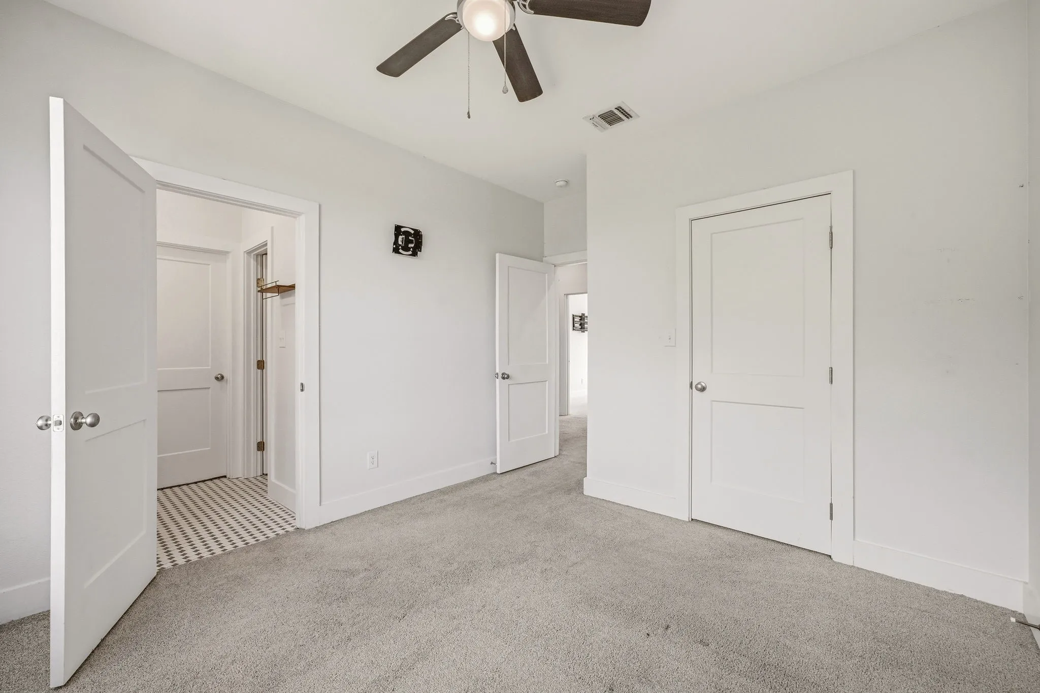 Light gray carpeting with white baseboards, a ceiling fan with dark wood-finish blades, white two-panel interior doors with brushed nickel hardware, and a closet with a patterned tile floor