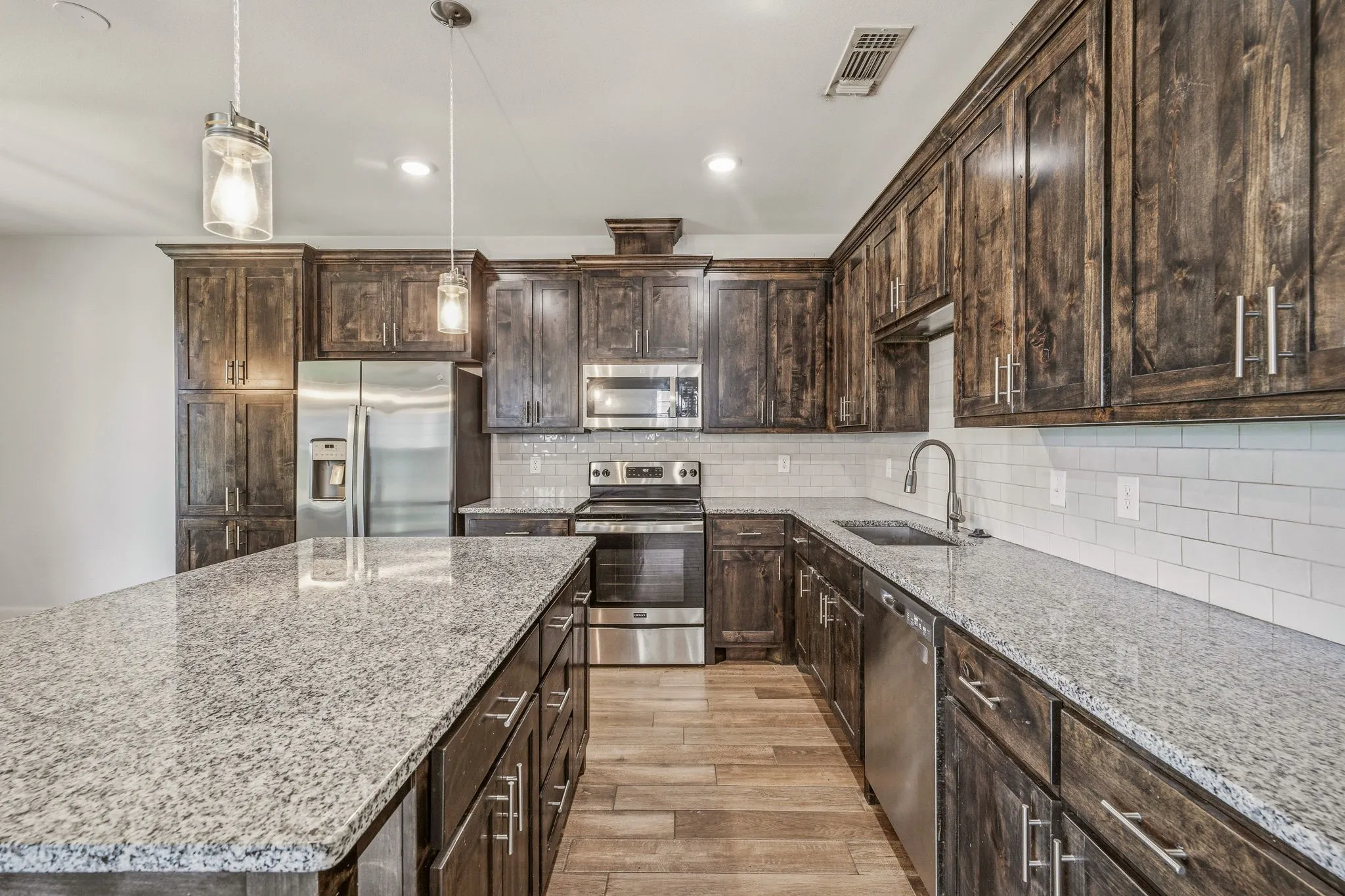 Kitchen featuring a large central island with light-toned countertops, rich wood-finish cabinetry, stainless steel appliances, a white subway tile backsplash, and wood-finish flooring