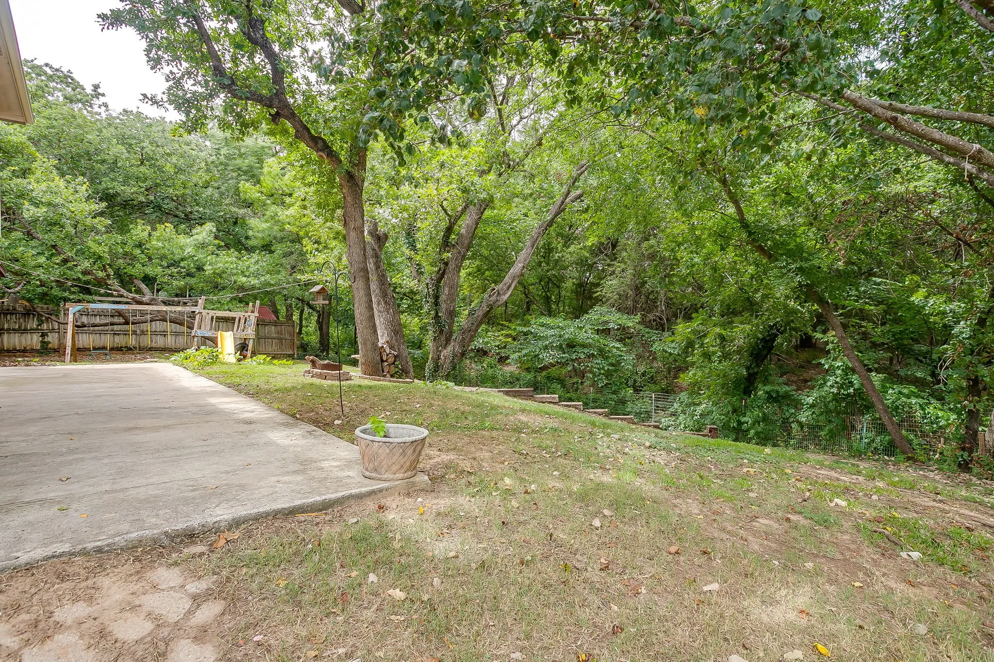 Expansive rear property featuring concrete patio, sloped grassy area, mature trees, wood privacy fencing, and terraced landscaping
