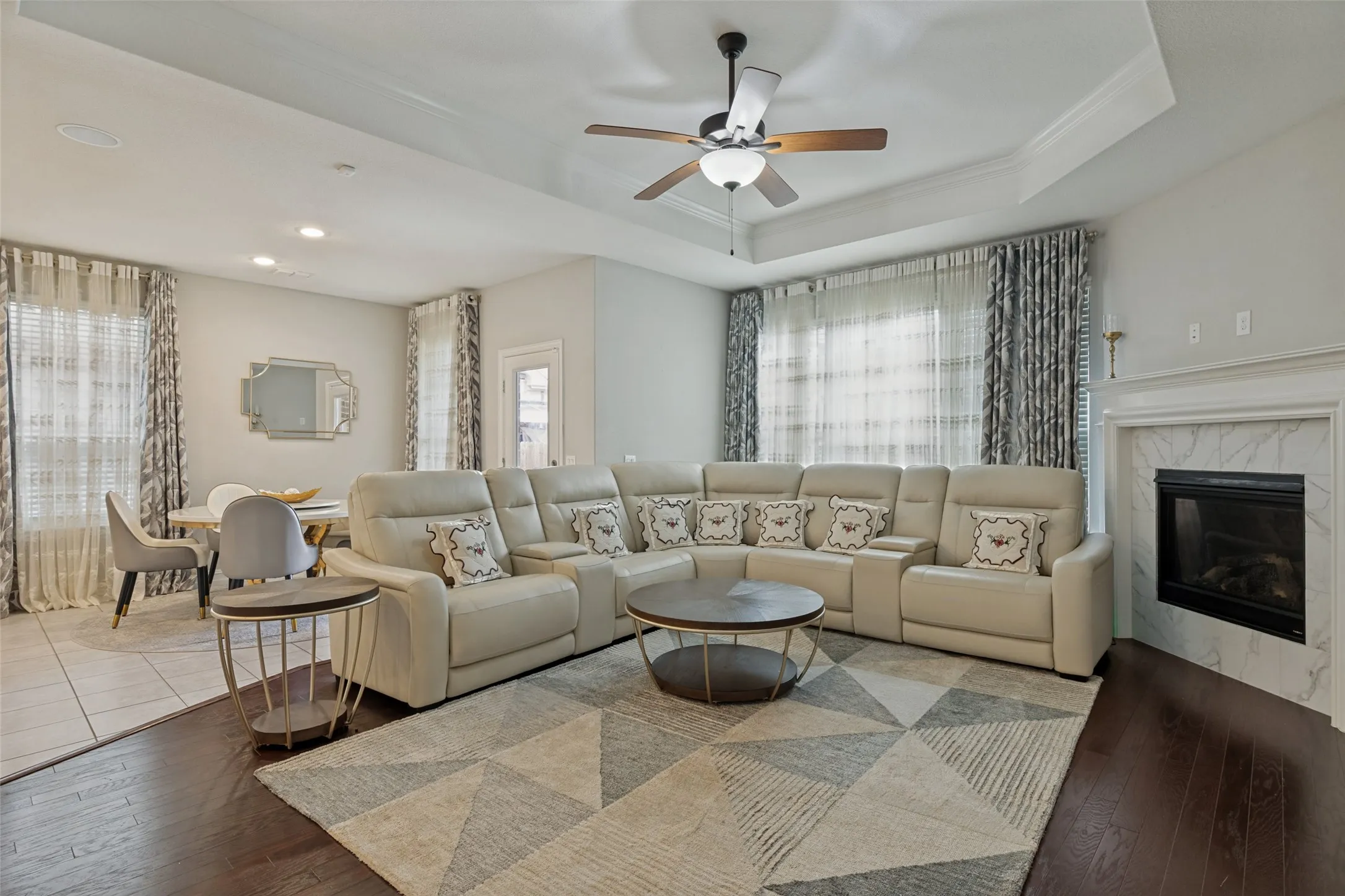 Open-concept living area featuring dark wood-finish flooring, a tray ceiling with a ceiling fan, recessed lighting, and a fireplace with a white mantel and marble-look surround