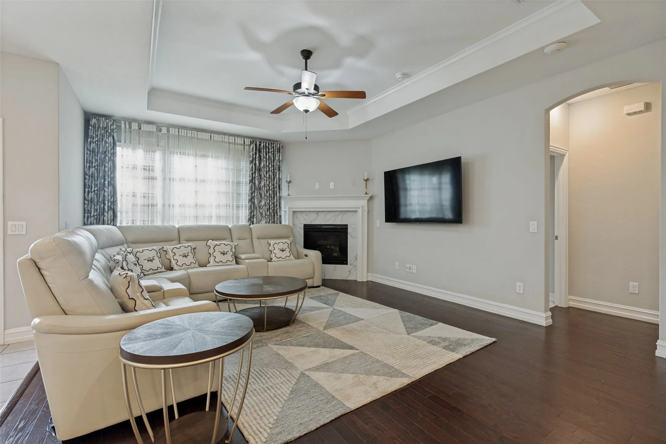 Spacious living area featuring dark wood-finish flooring, a tray ceiling with a ceiling fan, and a fireplace with a white mantel and dark stone surround
