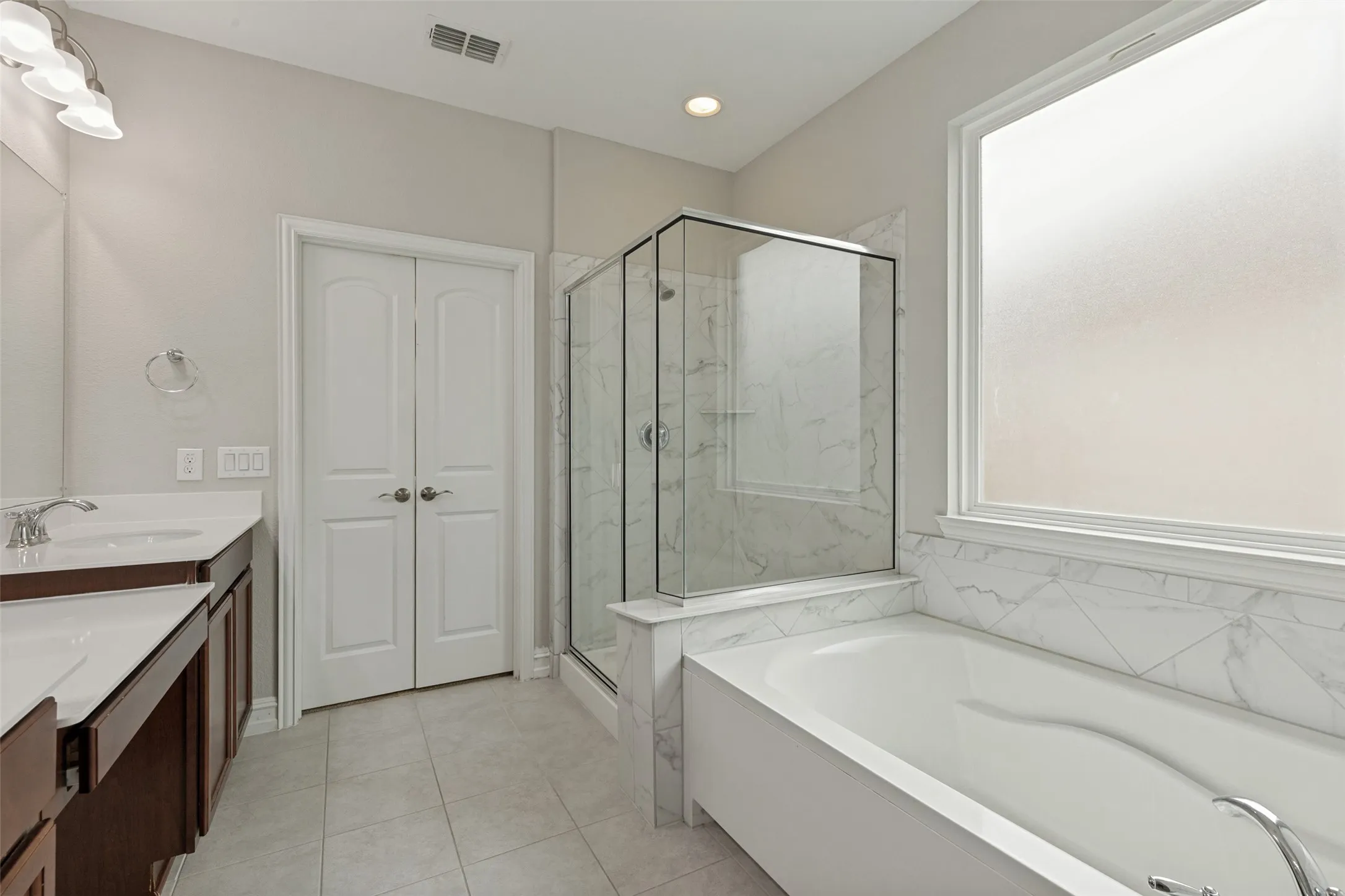 Contemporary bathroom featuring a large soaking tub with marble-patterned tile surround, a glass-enclosed shower, a double vanity with white countertops, and light gray floor tiling