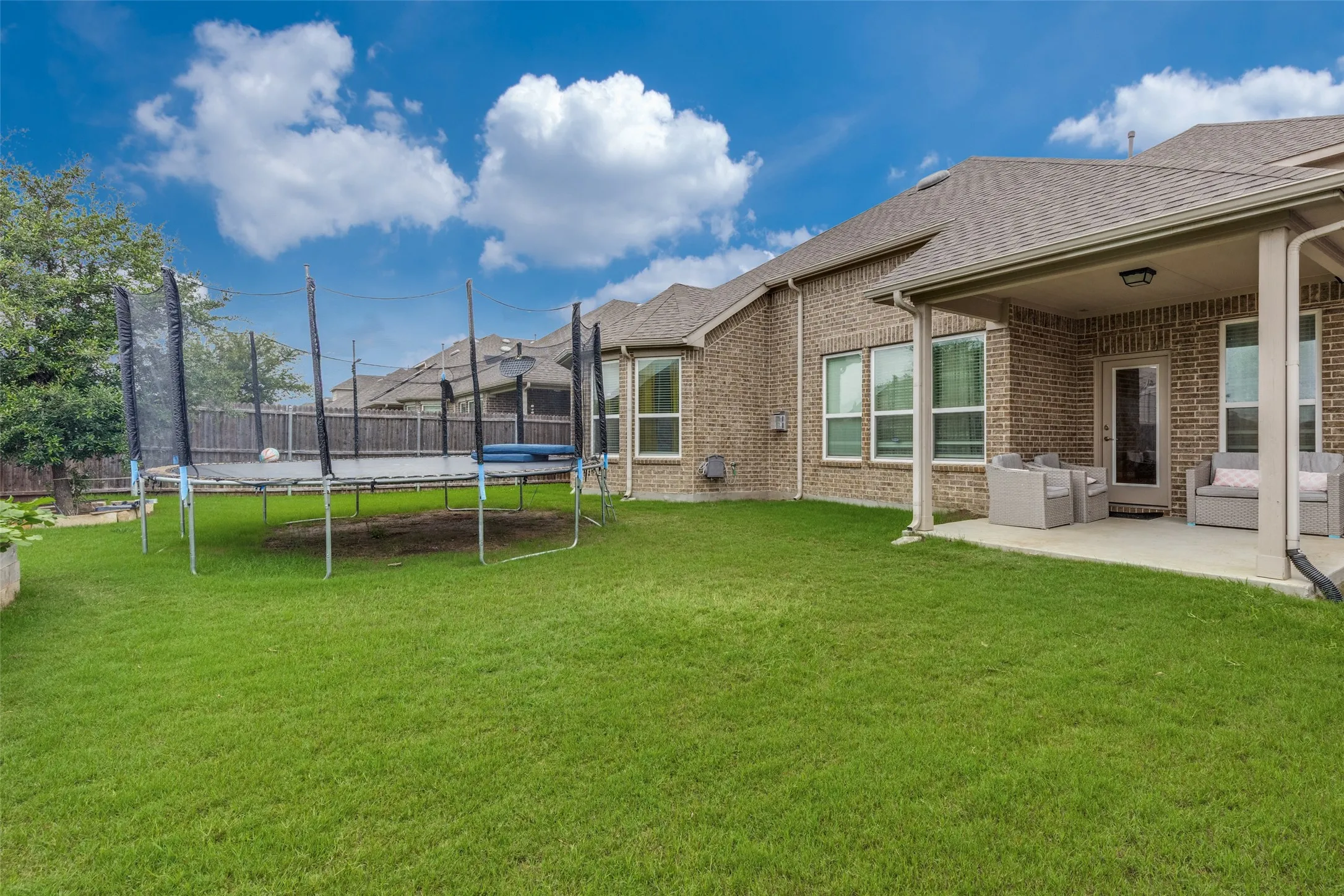 Expansive backyard featuring a manicured lawn, brick exterior, covered patio with concrete flooring, multiple large windows, and a privacy fence