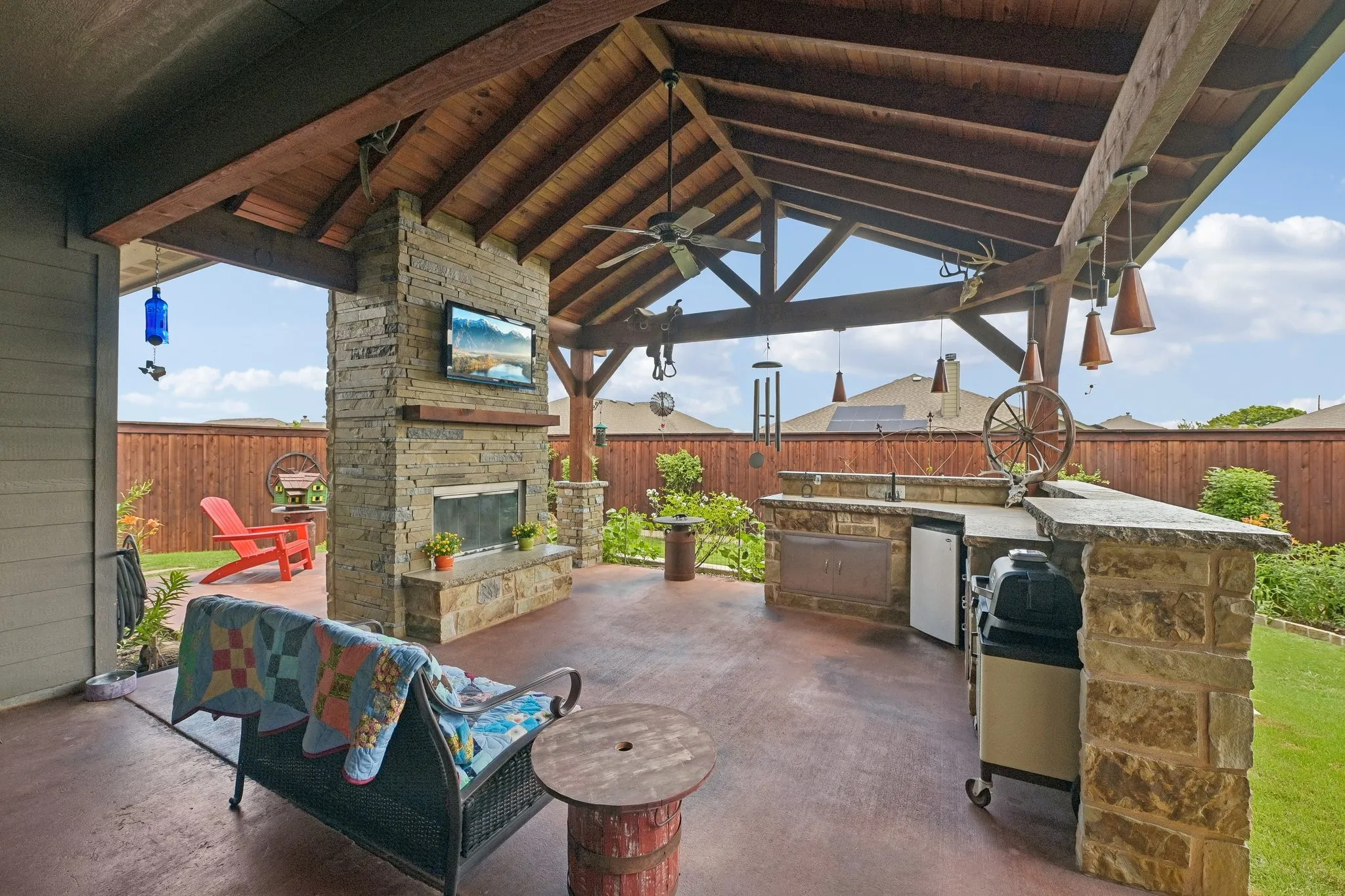 Covered outdoor patio featuring a vaulted wood-finish ceiling, a stone-clad fireplace with a built-in television, a stone-clad outdoor kitchen with integrated appliances, pendant lighting, and a stained concrete patio surface