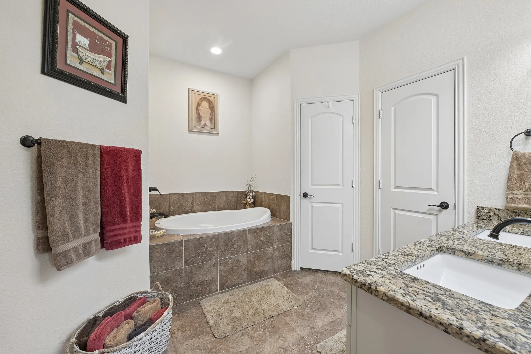 Soaking tub with tiled surround, dual white panel doors with black hardware, and a vanity featuring a granite-finish countertop, undermount sink, and dark-finish faucet