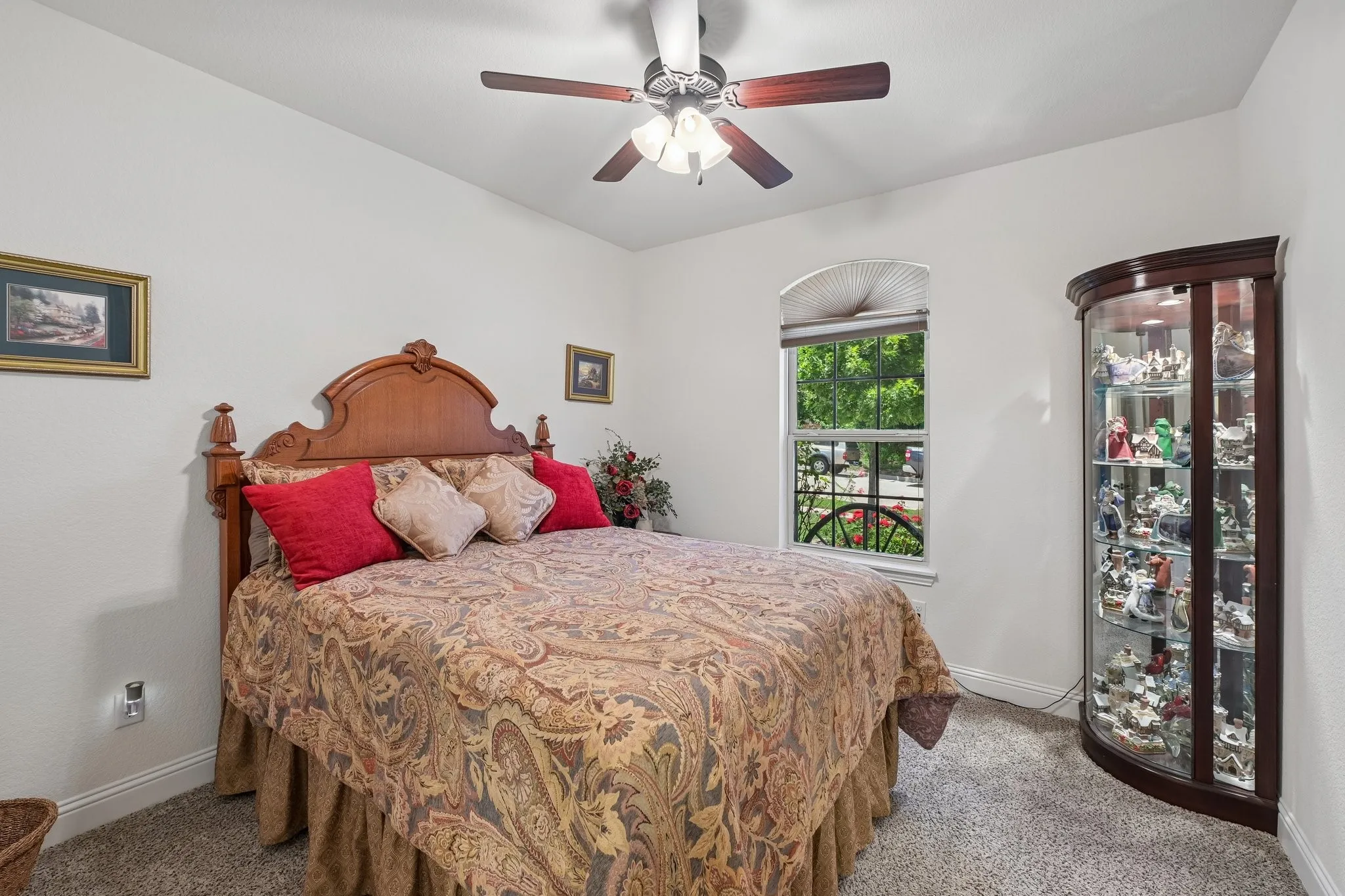 Carpeted bedroom featuring a ceiling fan with light fixture, a window with a valance, and a built-in corner display cabinet with glass shelving
