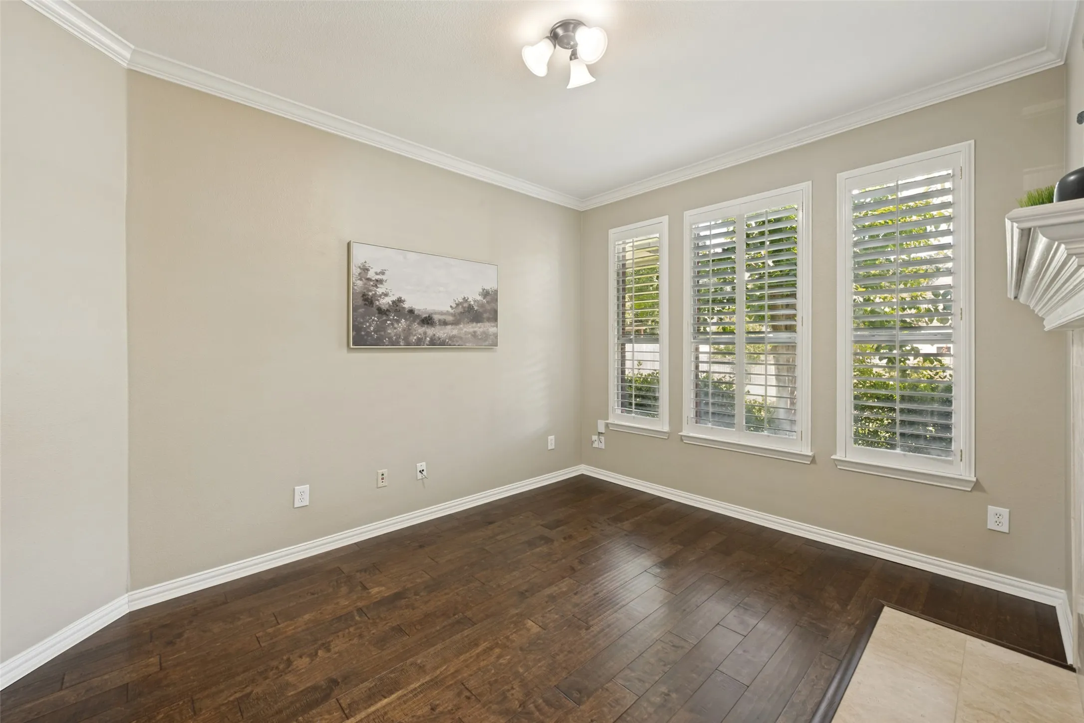 Spacious room featuring wood-finish flooring, three windows with plantation shutters, crown molding, and a built-in fireplace mantel