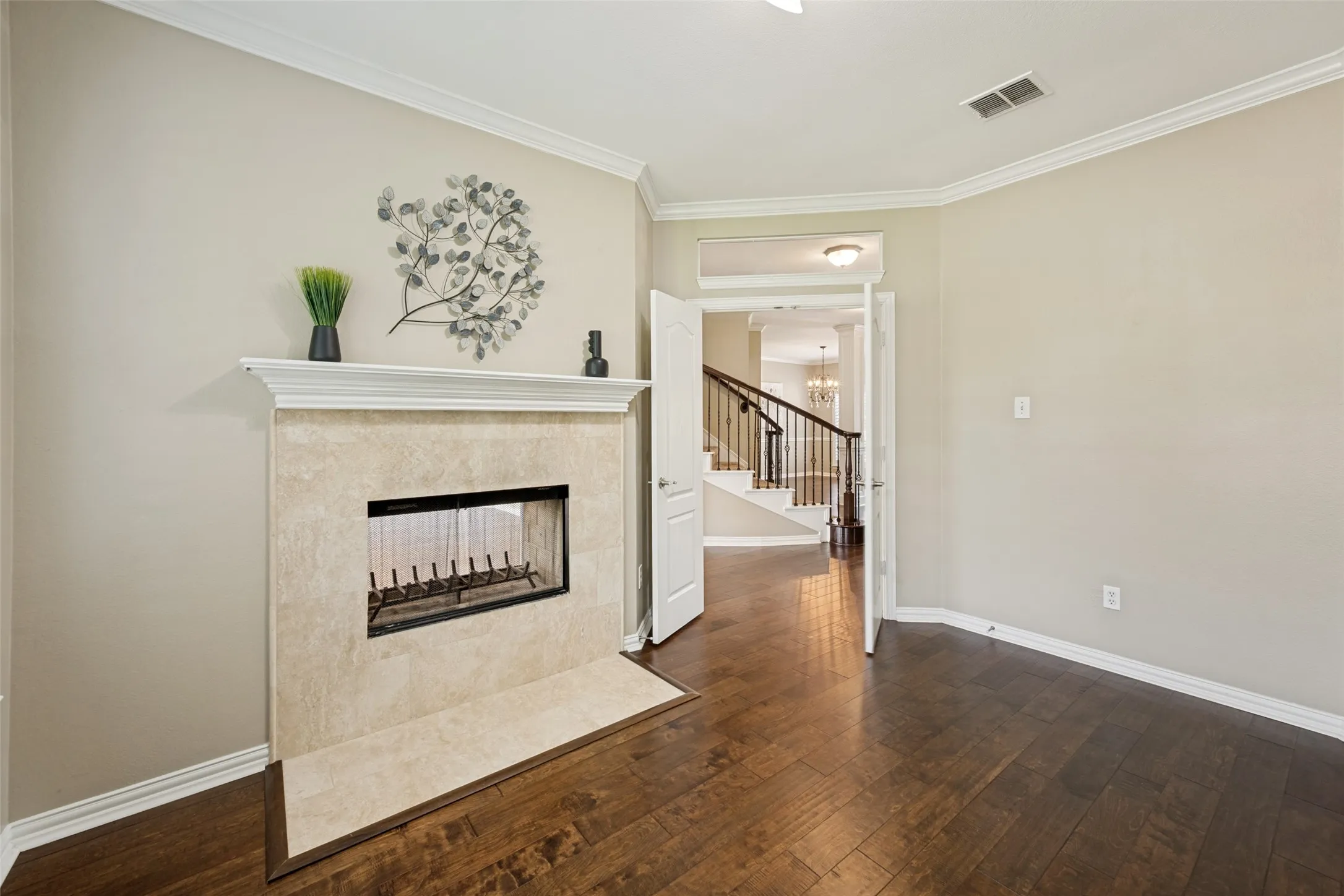 Living area featuring a floor-to-ceiling fireplace with a stone surround and mantel, rich wood-finish flooring, crisp white crown molding, and a visible staircase with dark wood handrails and white balusters