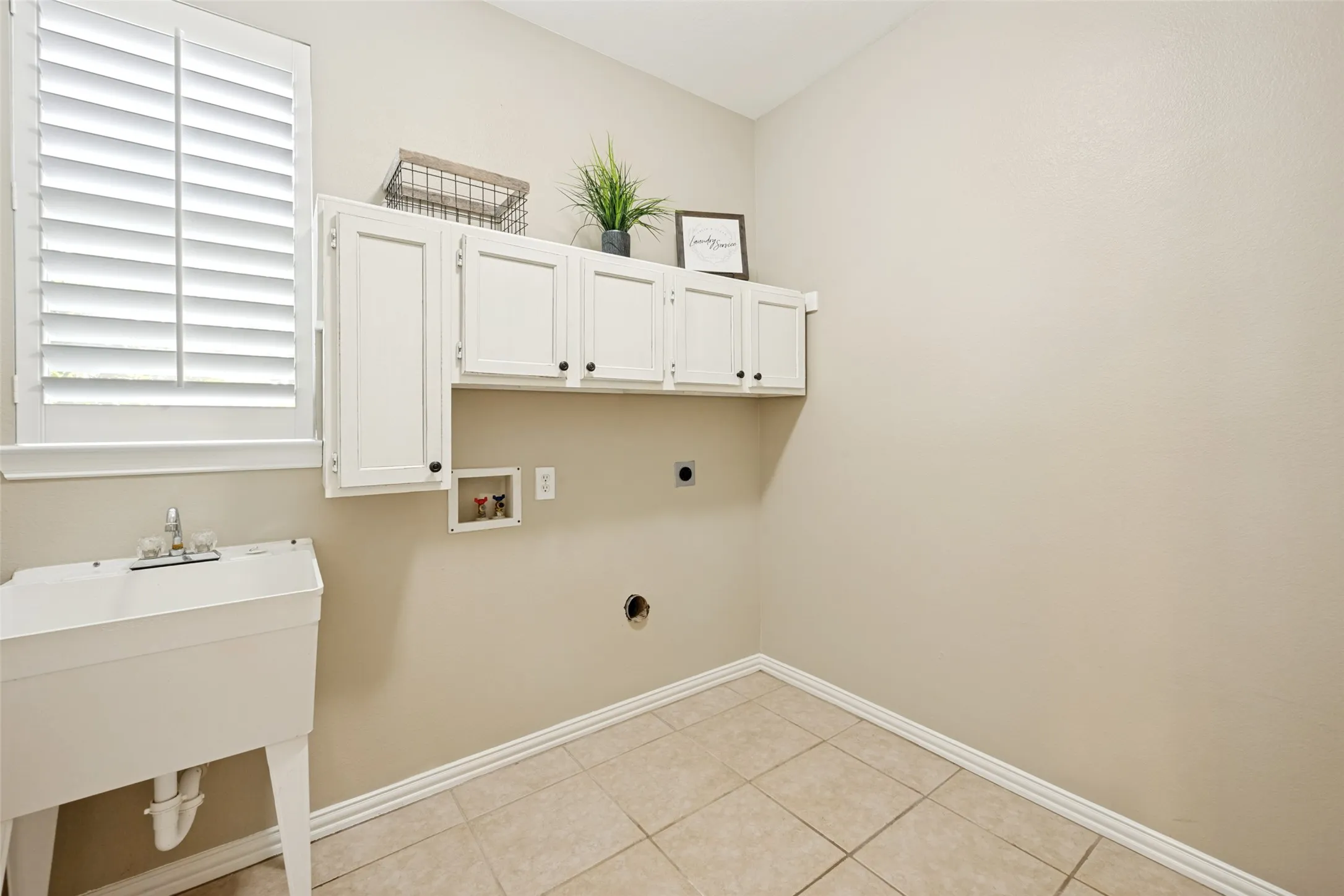 Utility room featuring a built-in wash basin, white wall-mounted cabinetry, window with plantation shutters, tile flooring, and neutral wall tones