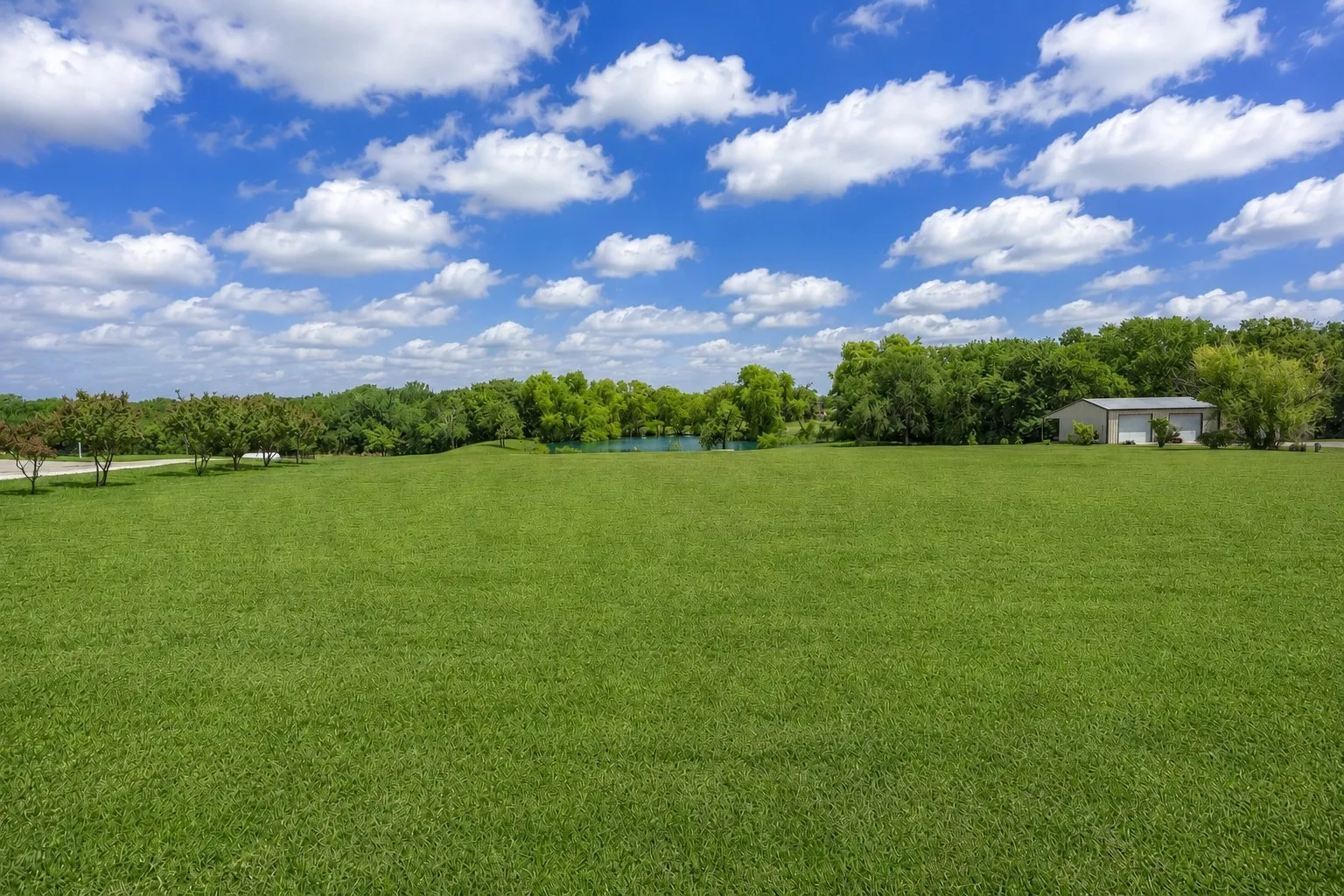 Expansive green space featuring a large metal building with multiple garage doors, mature trees, and a visible body of water