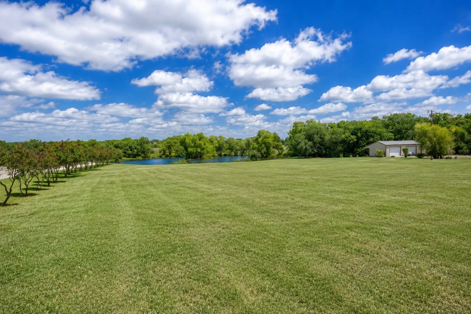 Expansive green lawn with a tree-lined perimeter, a visible body of water, and a two-door outbuilding