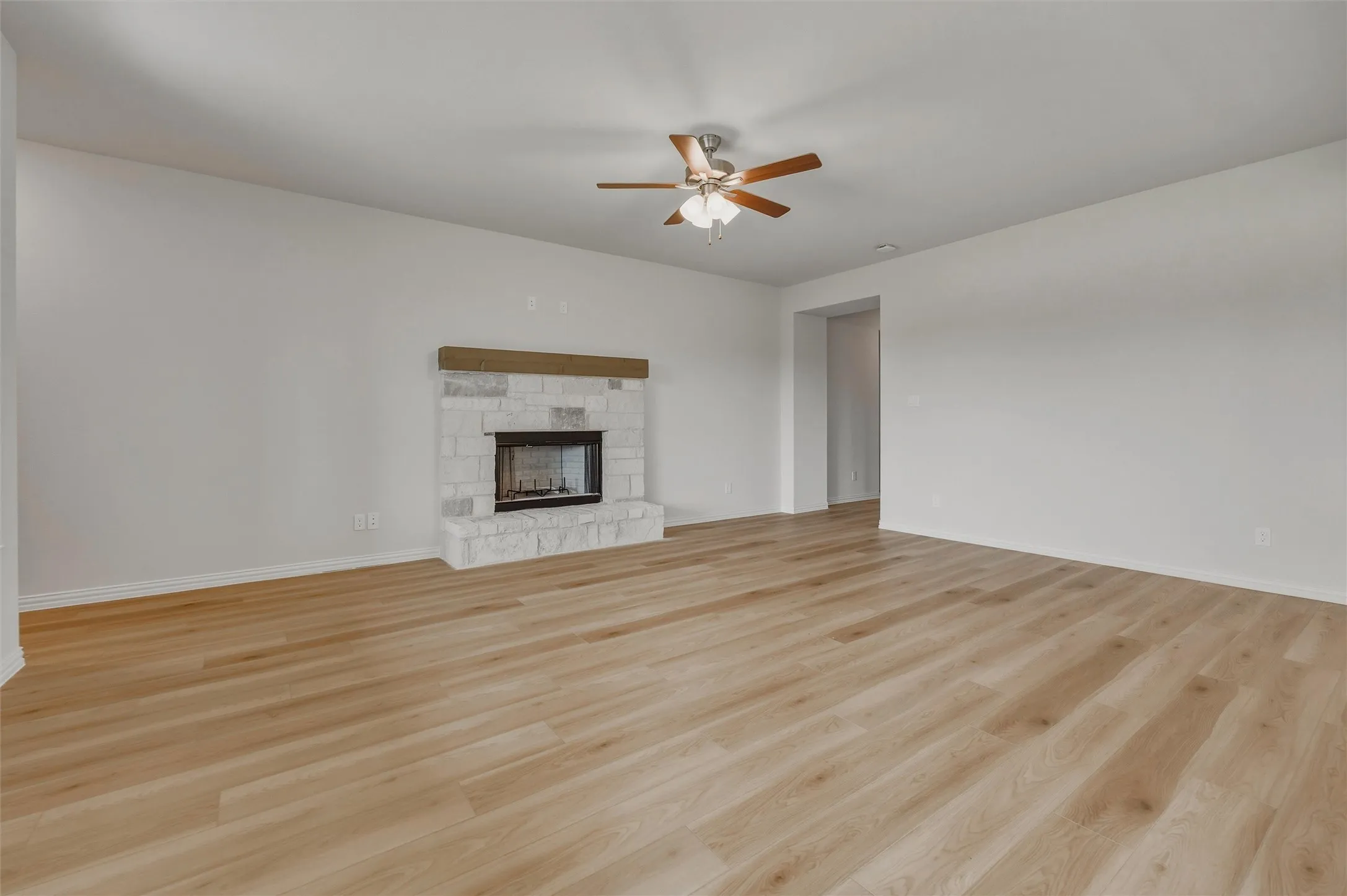 Spacious interior with light-tone wood-finish flooring, a white stone-clad fireplace with a wood mantel, and a ceiling fan