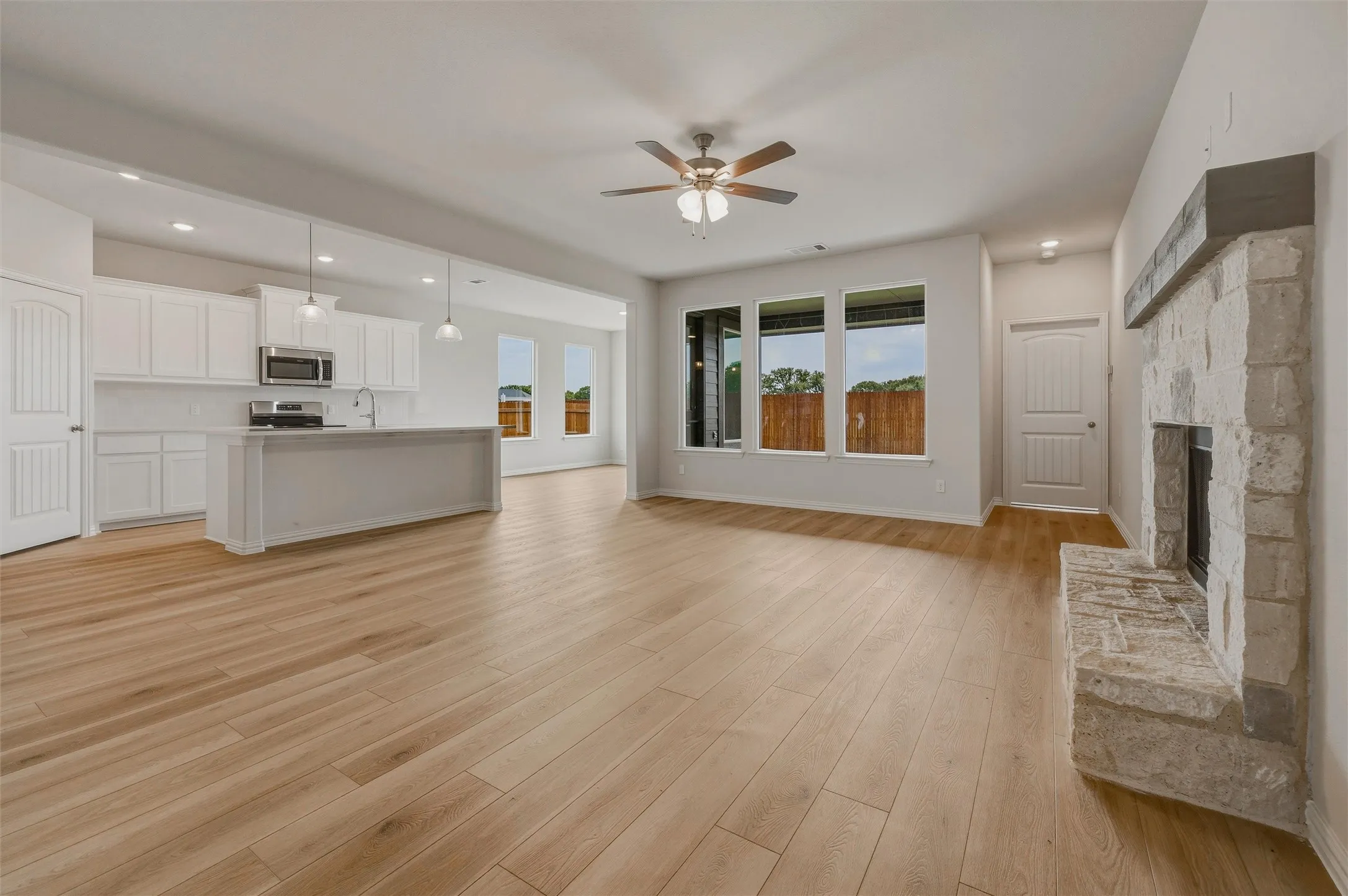 Wood-finish flooring, stone-clad fireplace with mantel, white kitchen cabinetry, stainless steel appliances, and a kitchen island with pendant lighting